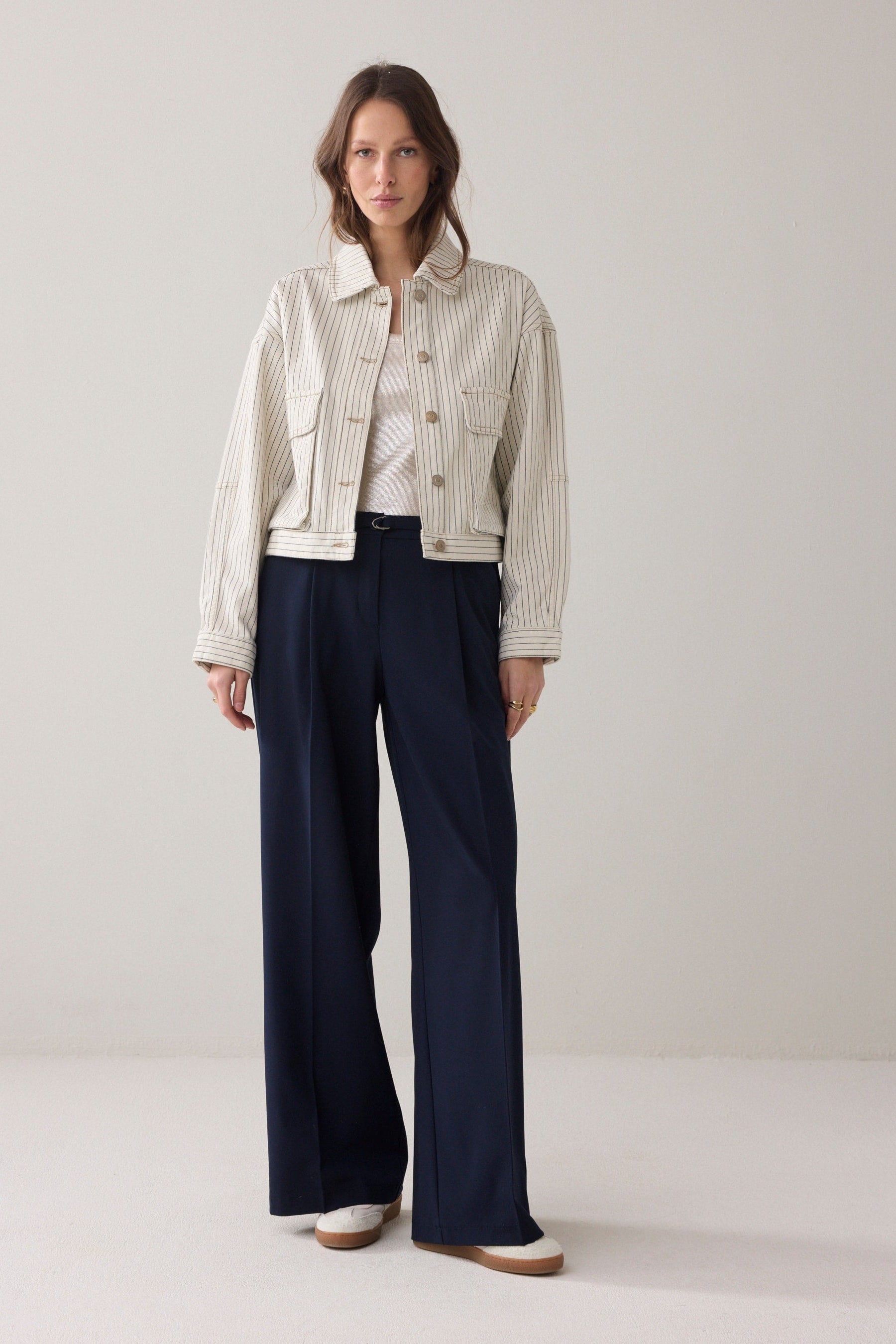 A women wearing cropped denim jacket wth an ivory base and navy herringbone vertical stripe detail  and navy wide leg paints and light coloured sneakers on a plain background.  