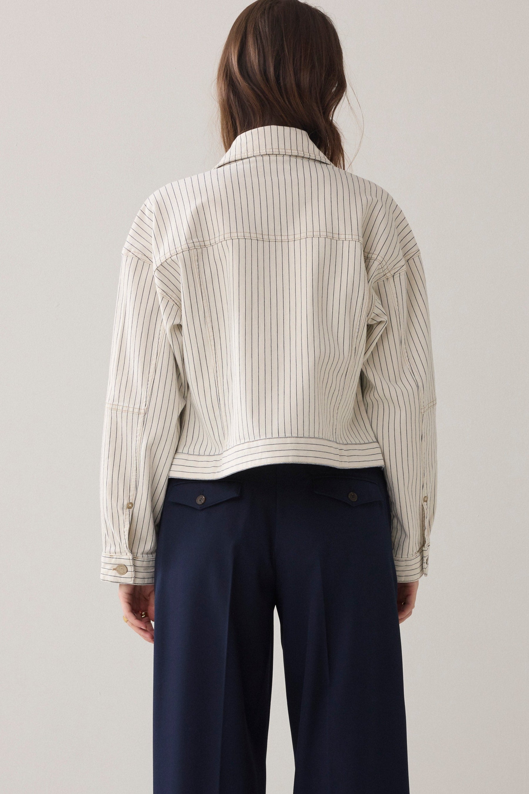 A women shown from the back wearing cropped denim jacket wth an ivory base and navy herringbone vertical stripe detail  and navy wide leg paints on a plain background.  