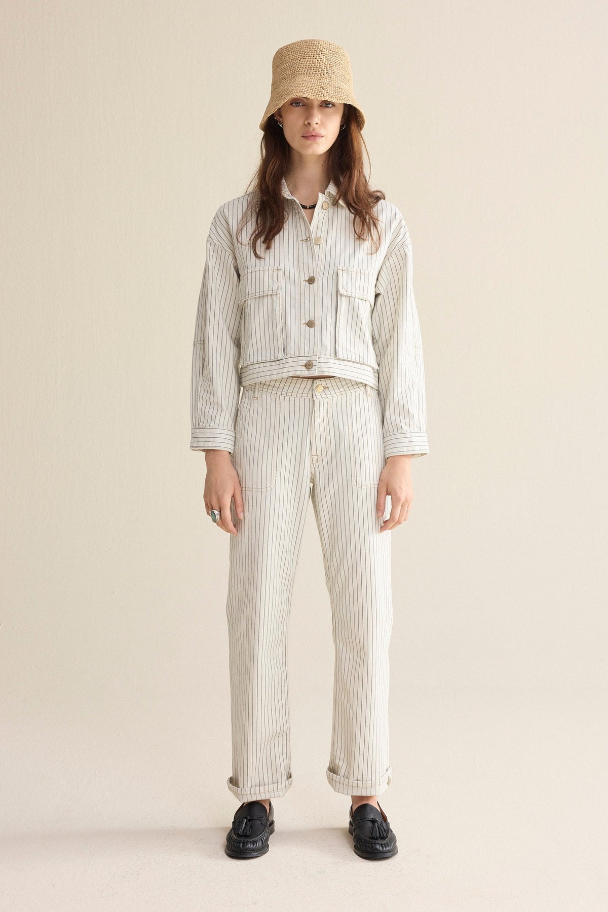 Model wearing Ivory Denim pants with horizontal navy herringbone stripe and gold domes with a matching jacket with black loafers and a straw bucket hat on a plain background. 