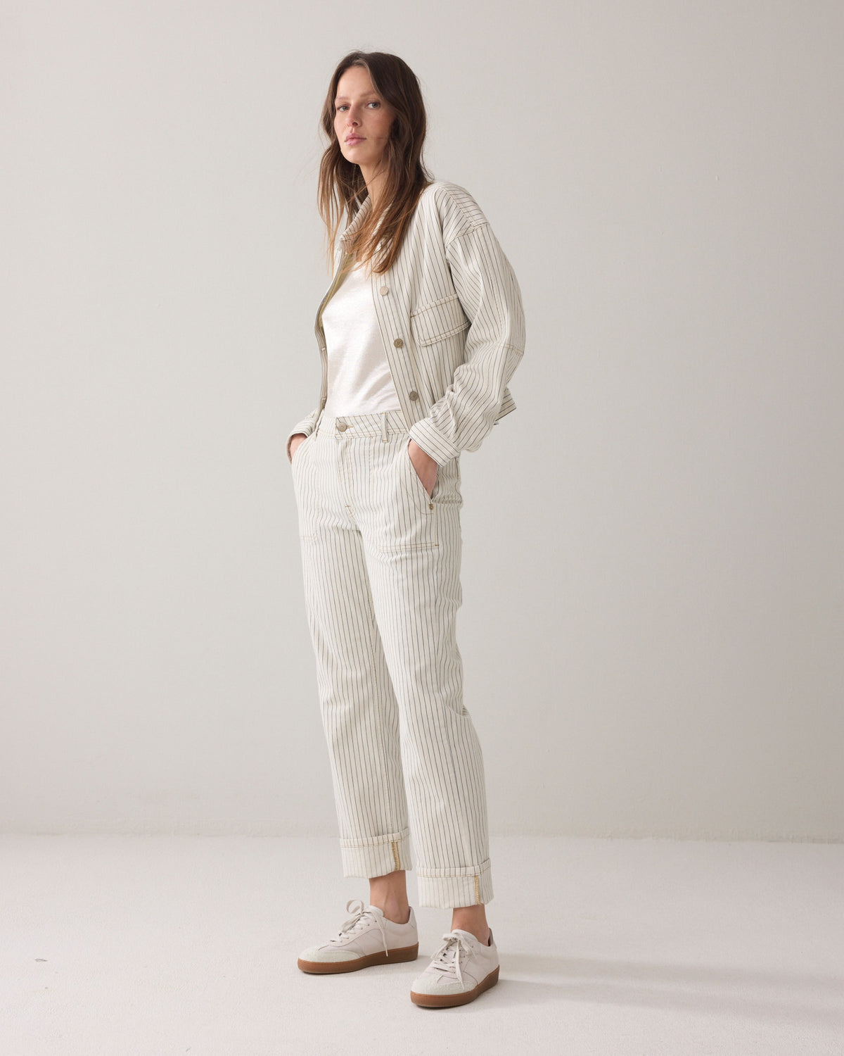Model wearing Ivory Denim pants with horizontal navy herringbone stripe and gold domes with a matching jacket over her shoulders and light coloured sneakers on a plain background. 