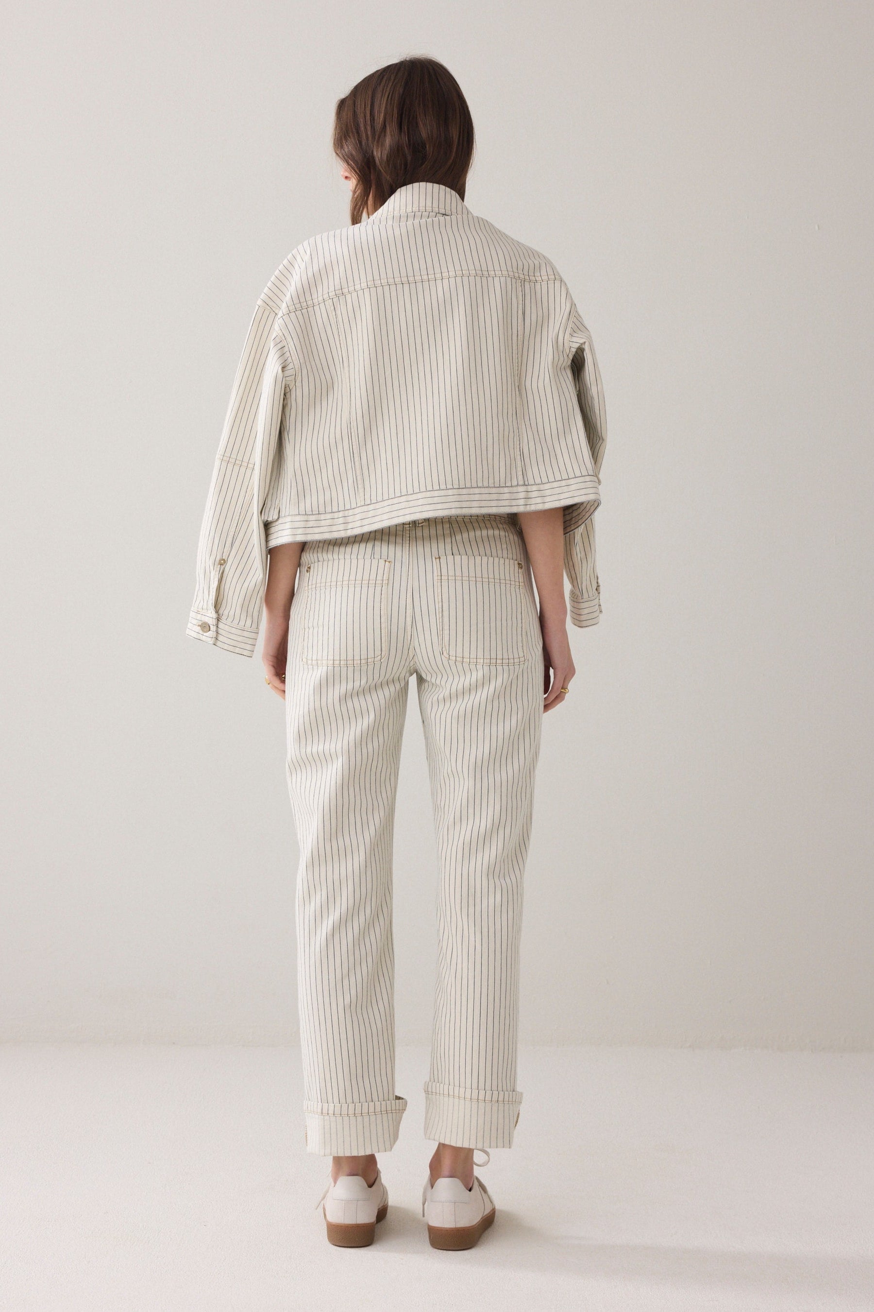 Model shown from the back wearing Ivory Denim pants with horizontal navy herringbone stripe and gold domes with a matching jacket over her shoulders and light coloured sneakers on a plain background. 