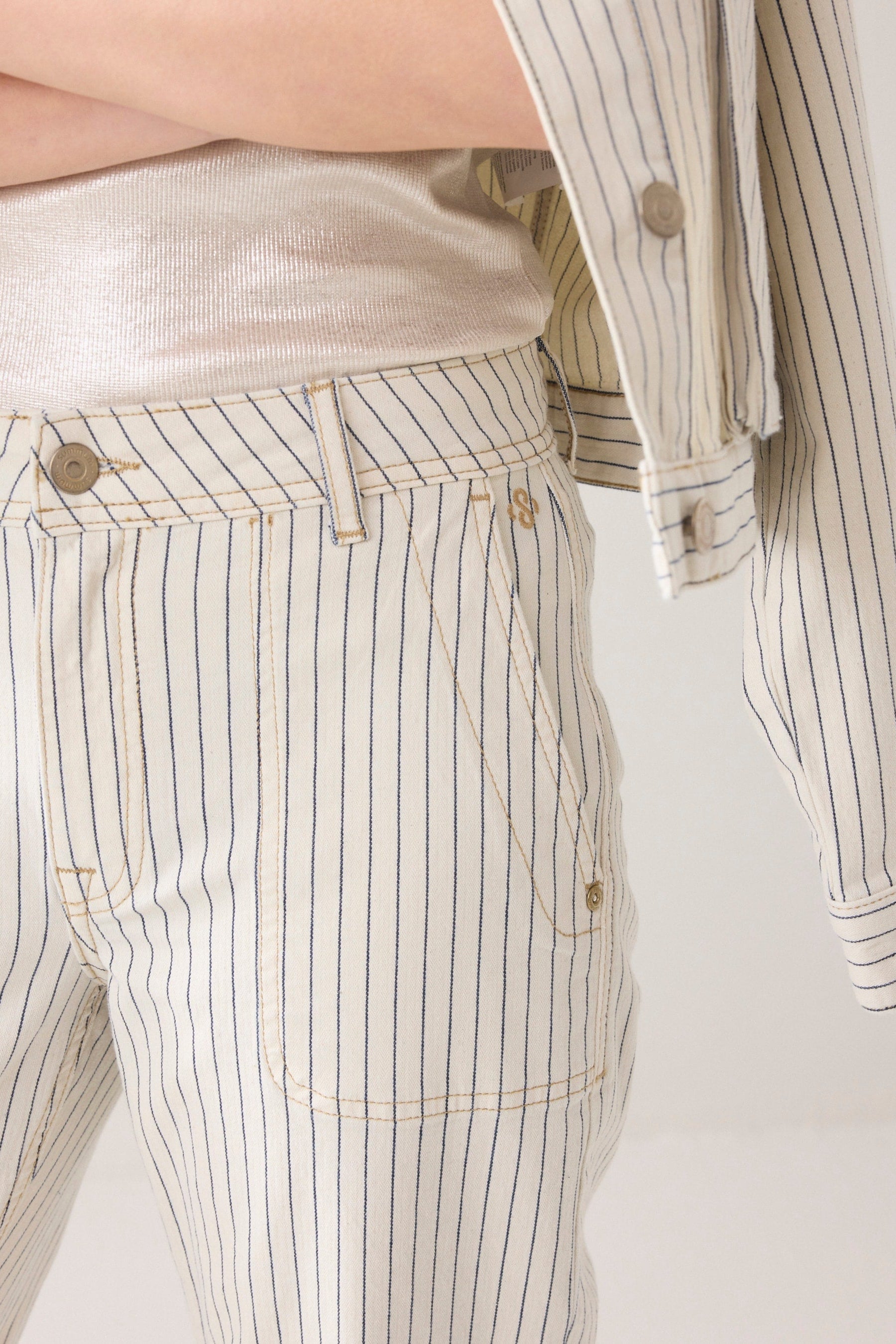Close up of model wearing Ivory Denim pants with horizontal navy herringbone stripe and gold domes on a plain background. 