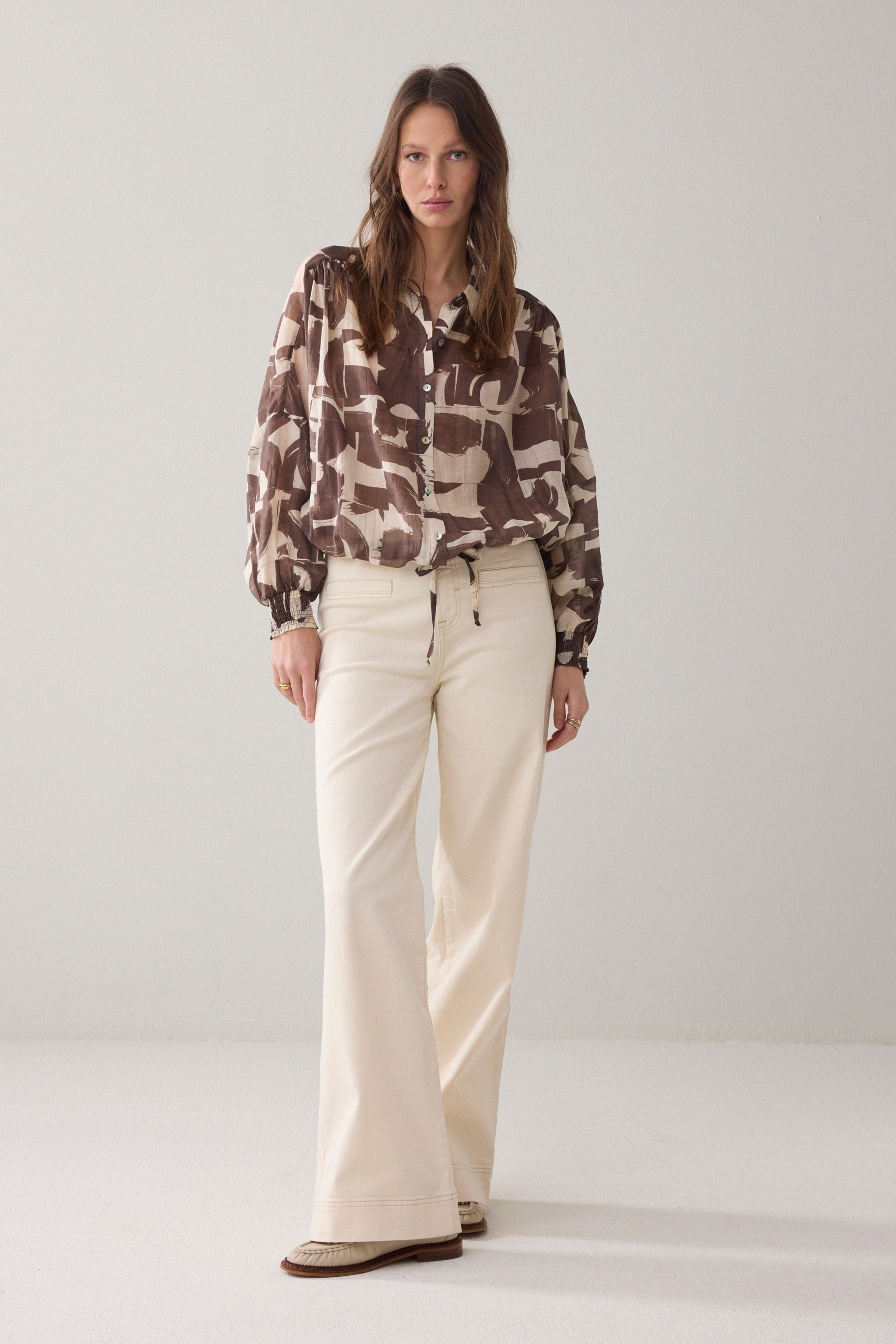Women wearing a brown and cream patterned  shirt and cream pants  on a plain background. 