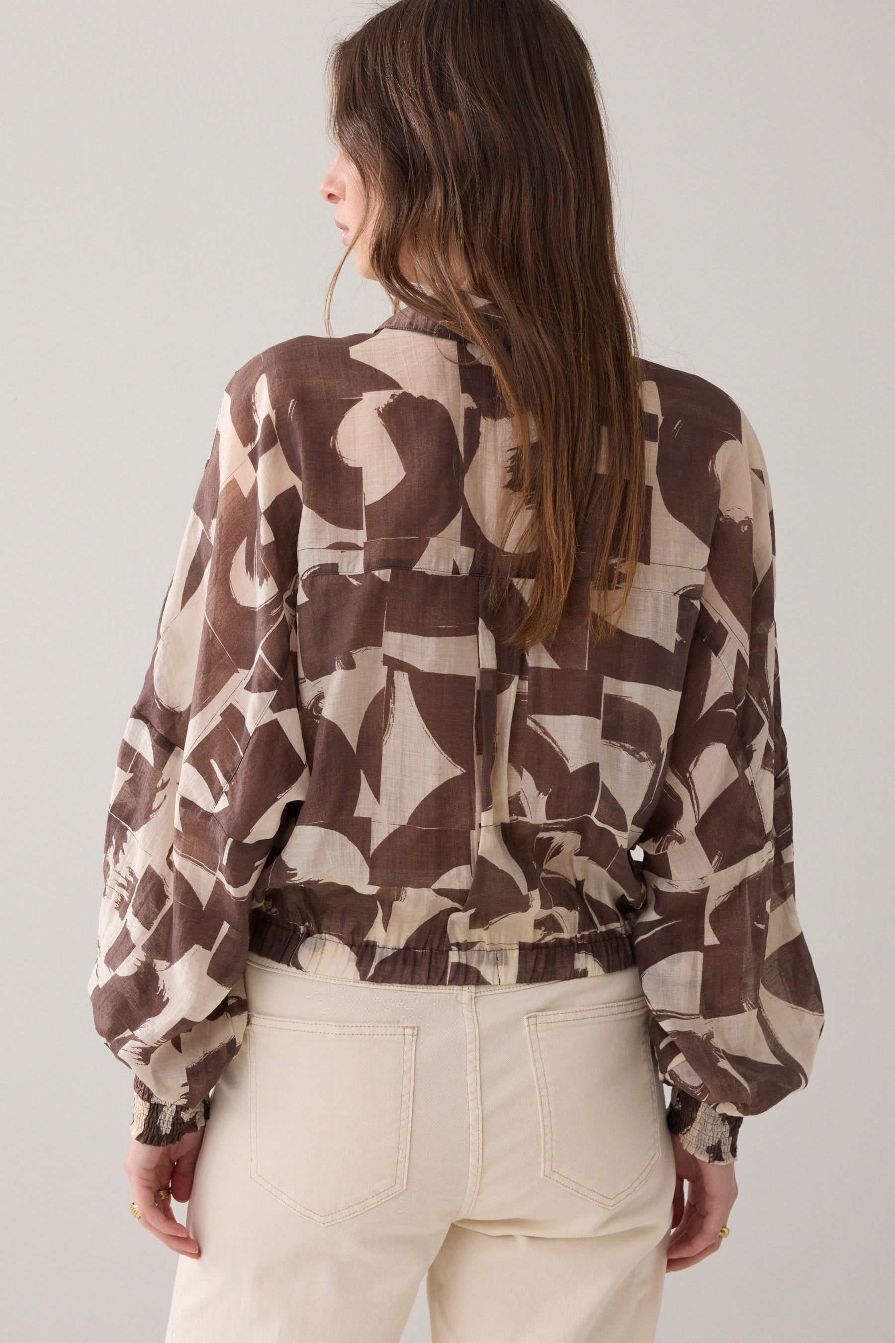Women wearing a brown and cream patterned  shirt and cream pants  on a plain background. 