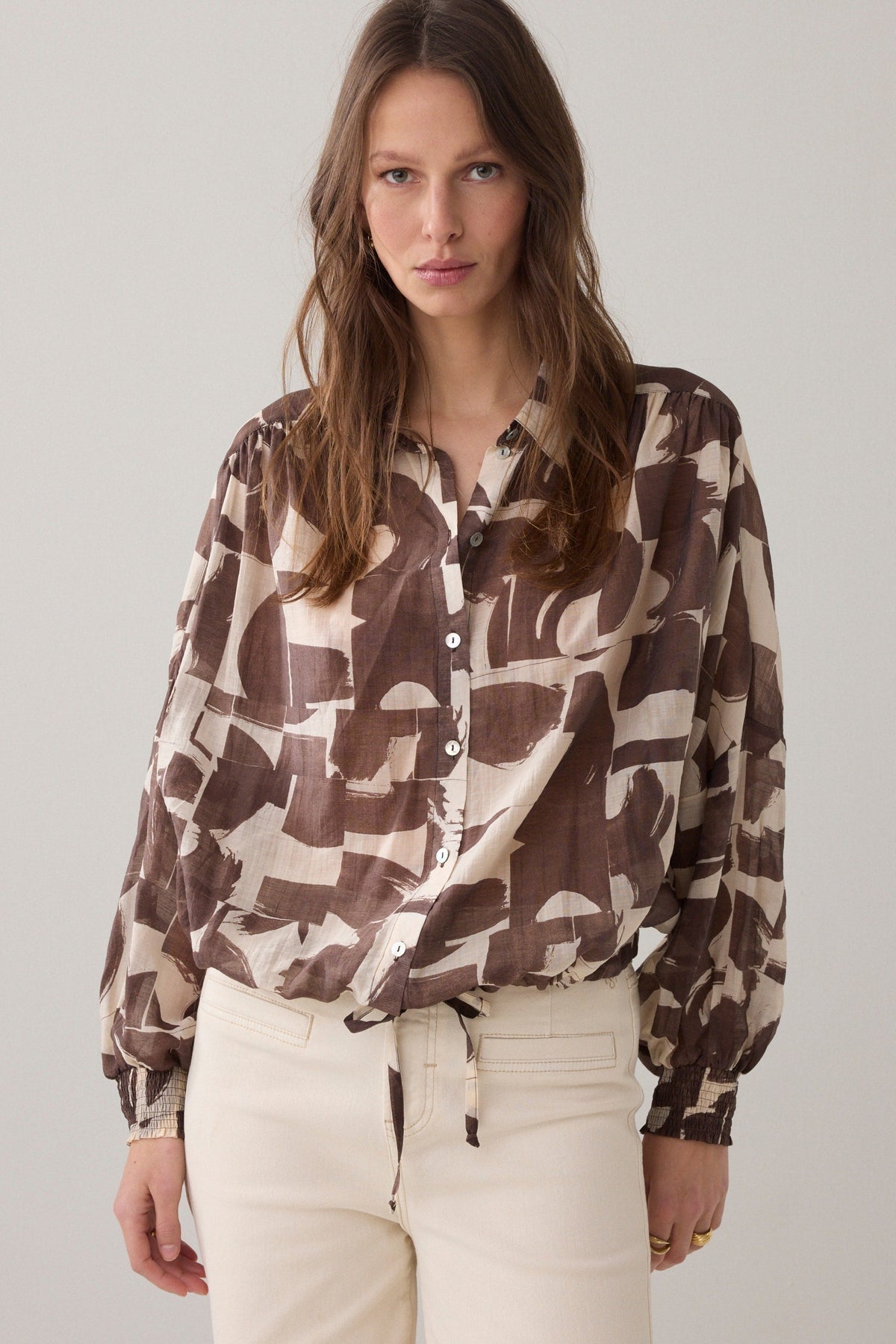 Women wearing a brown and cream patterned  shirt and cream pants  on a plain background. 