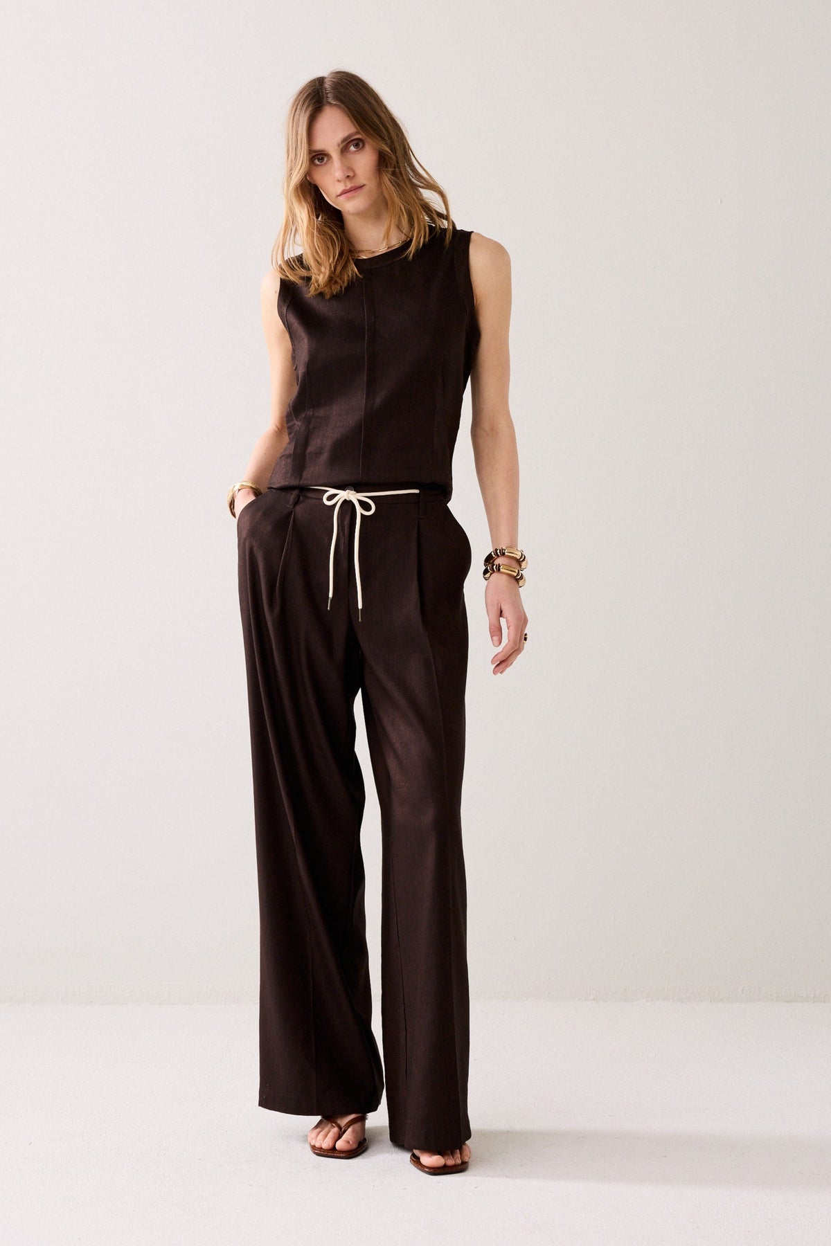 Woman shown from the back wearing wide leg dark brown trousers and a matching top with gold bracelets on a plain background. 