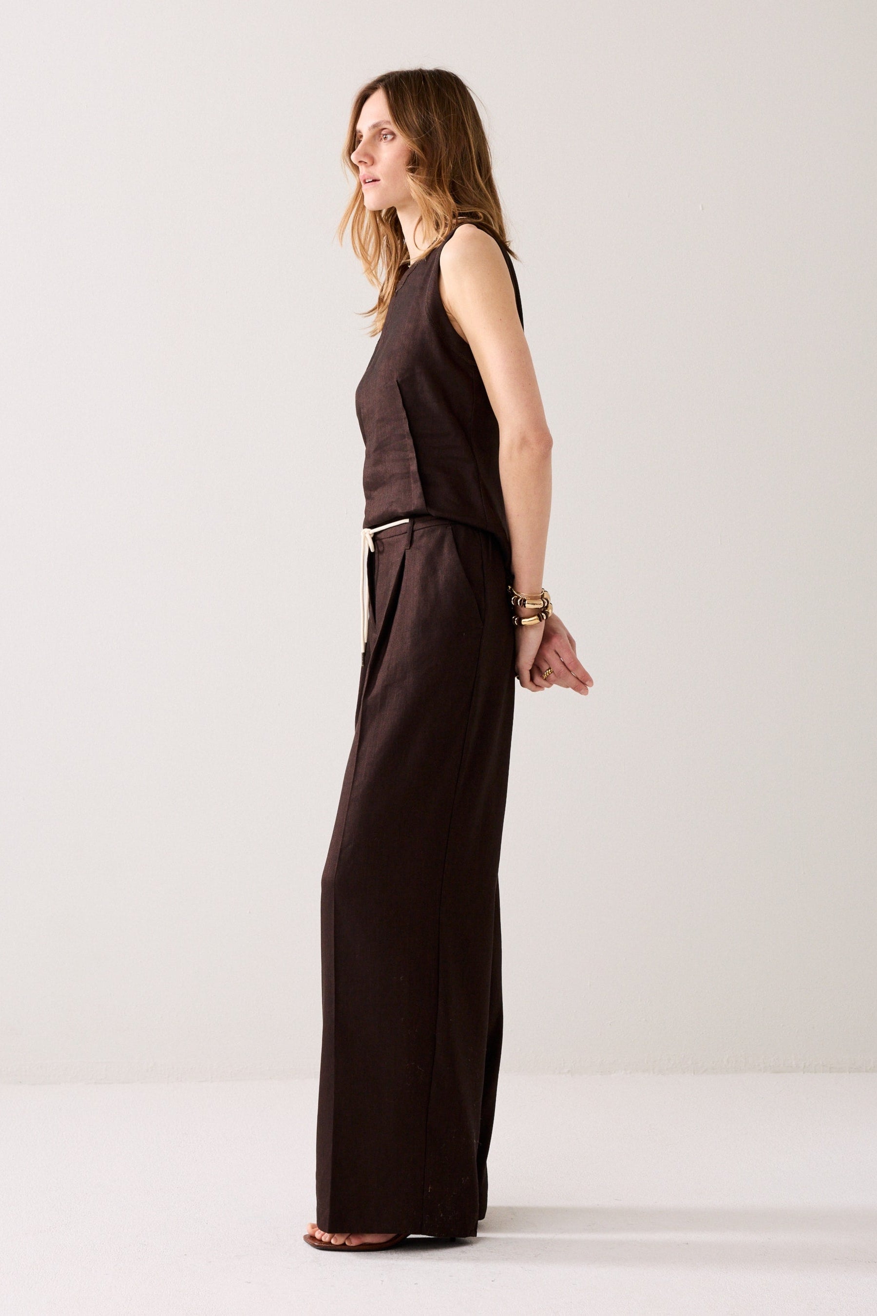 Woman shown from the side view wearing wide leg dark brown trousers and a matching top with gold bracelets on a plain background. 