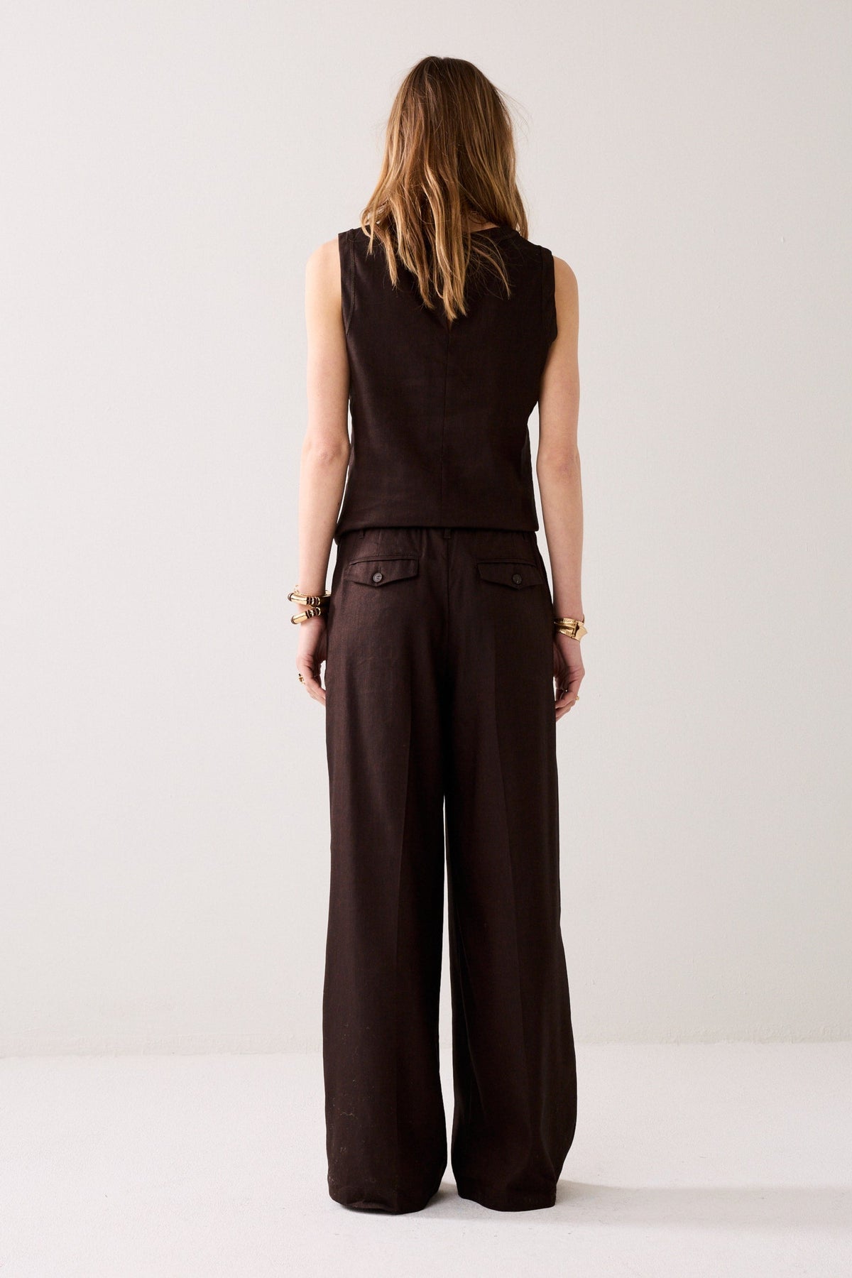 Woman shown from the back wearing wide leg dark brown trousers and a matching top with gold bracelets on a plain background. 