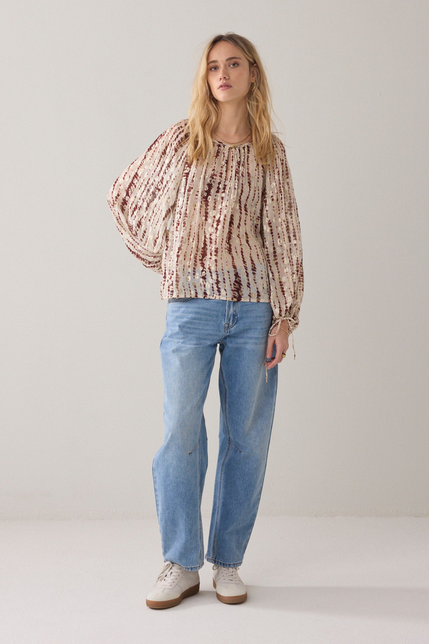 A woman wearing a coloured blouse with tie-dye pattern and voluminous balloon sleeves, paired with blue jeans against a light  background.