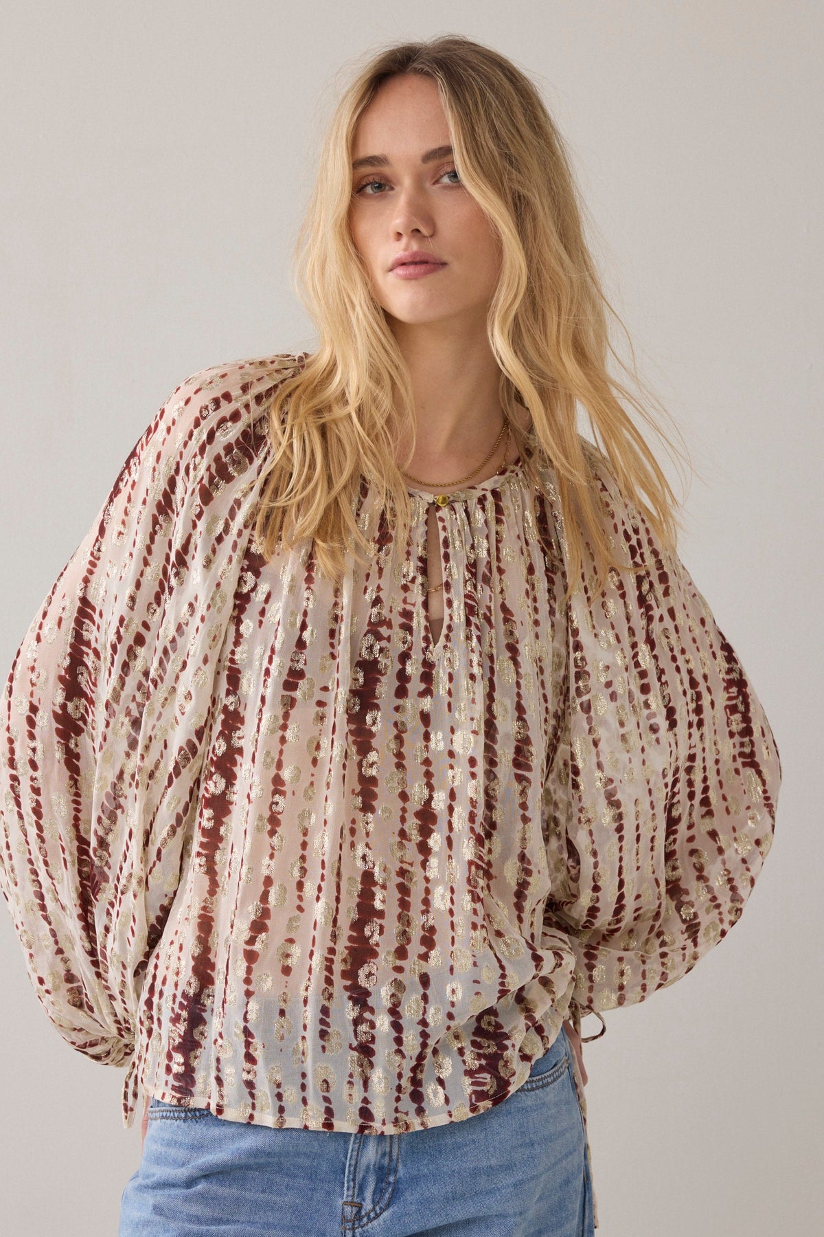 Woman wearing a tie-dye blouse with balloon sleeves and denim pants against a plain background