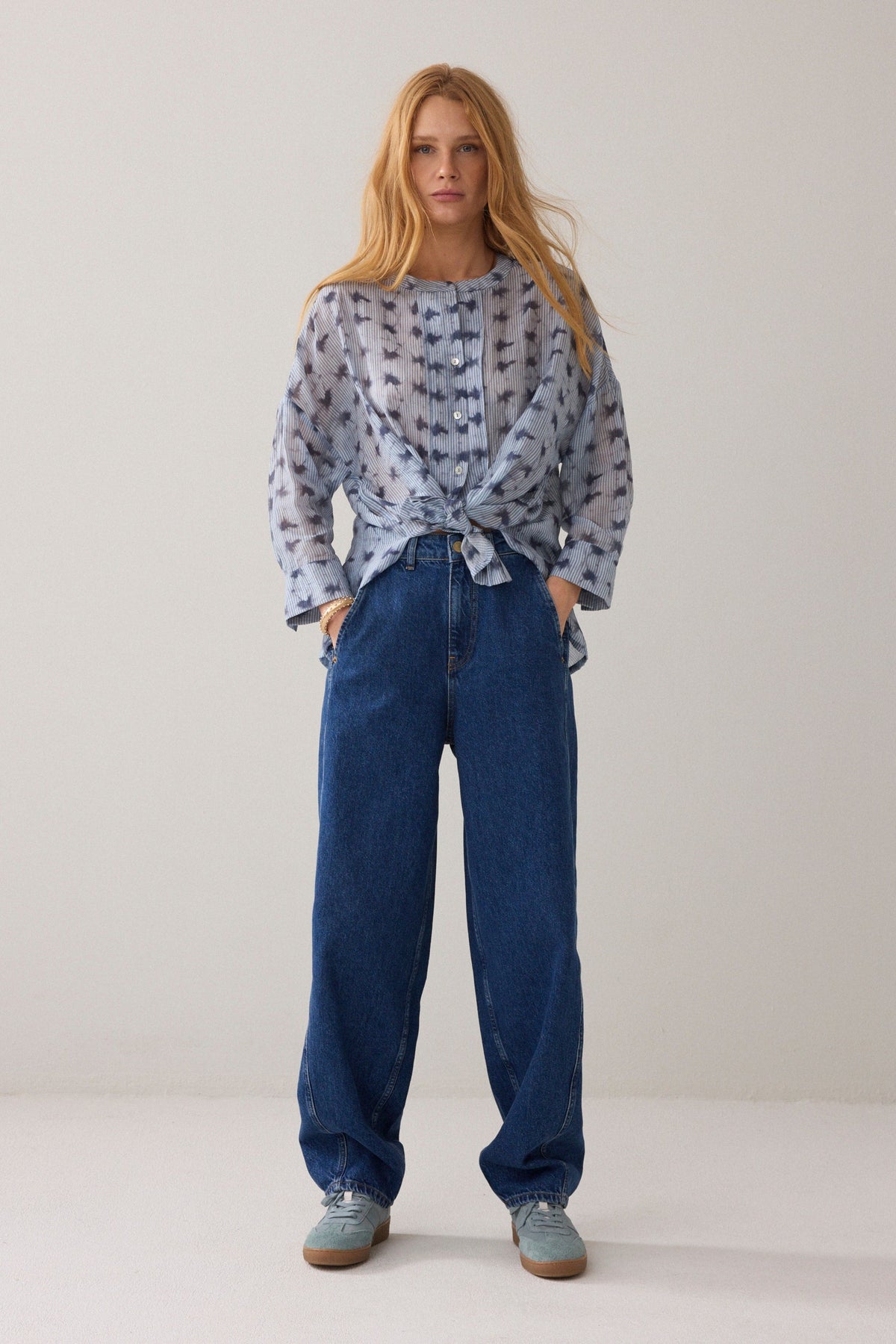 Women shown from the side wearing a light blue stripe pattern blouse with 3/4 sleeves and dark blue denim jeans and light blue sneakers on a plain background.