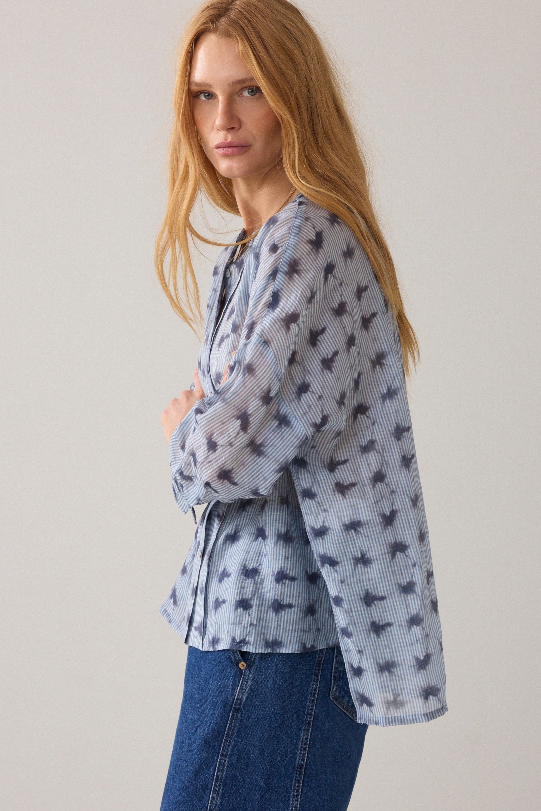Women shown from the side wearing a light blue stripe pattern blouse with 3/4 sleeves and dark blue denim jeans on a plain background. 