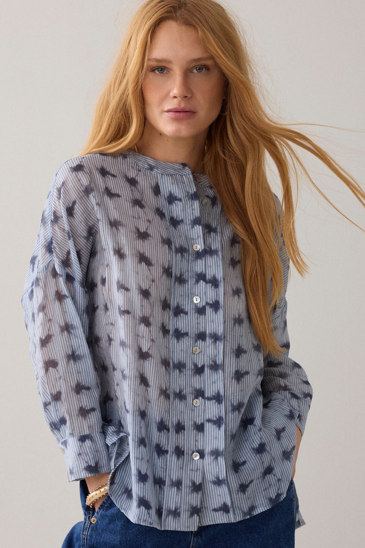 Women up close wearing a light blue stripe pattern blouse with 3/4 sleeves and dark blue denim jeans on a plain background.