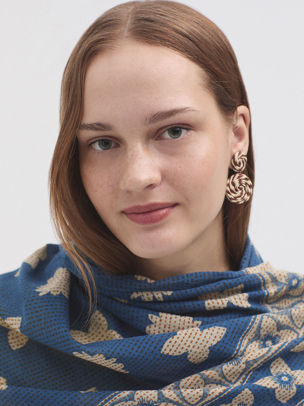 Woman wearing a blue patterned scarf and gold earrings against a white background