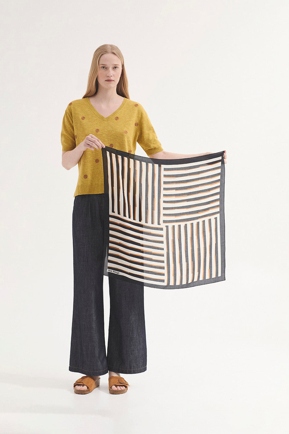 Model holding  scarf  in cream, navy and tan stripe wearing navy pants and a mustard spot top  on a plain background. 
