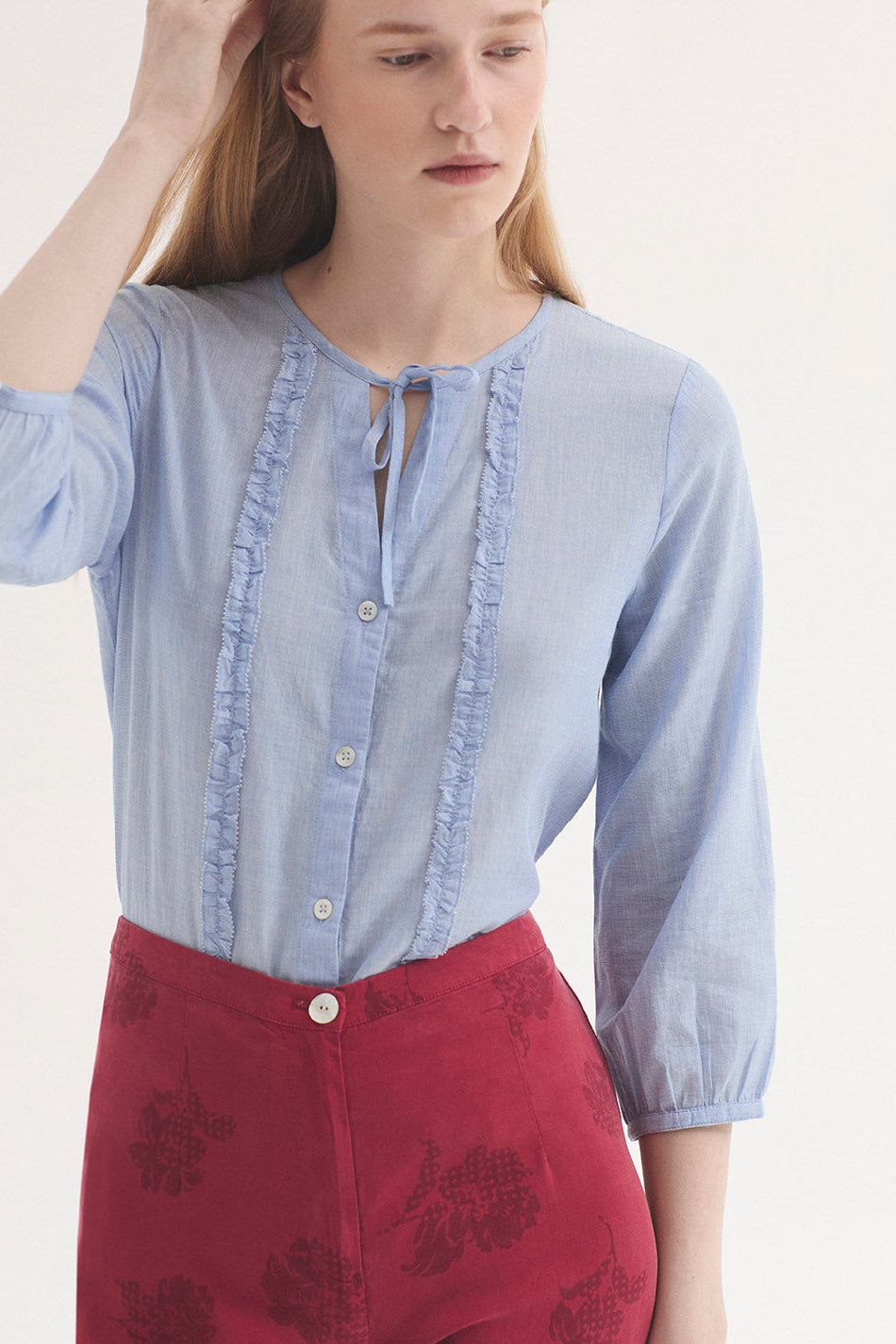 A woman wearing a light blue 3/4sleeve blouse with ruffle trim and a tie neckline, paired with red patterned trousers, against a plain light background.