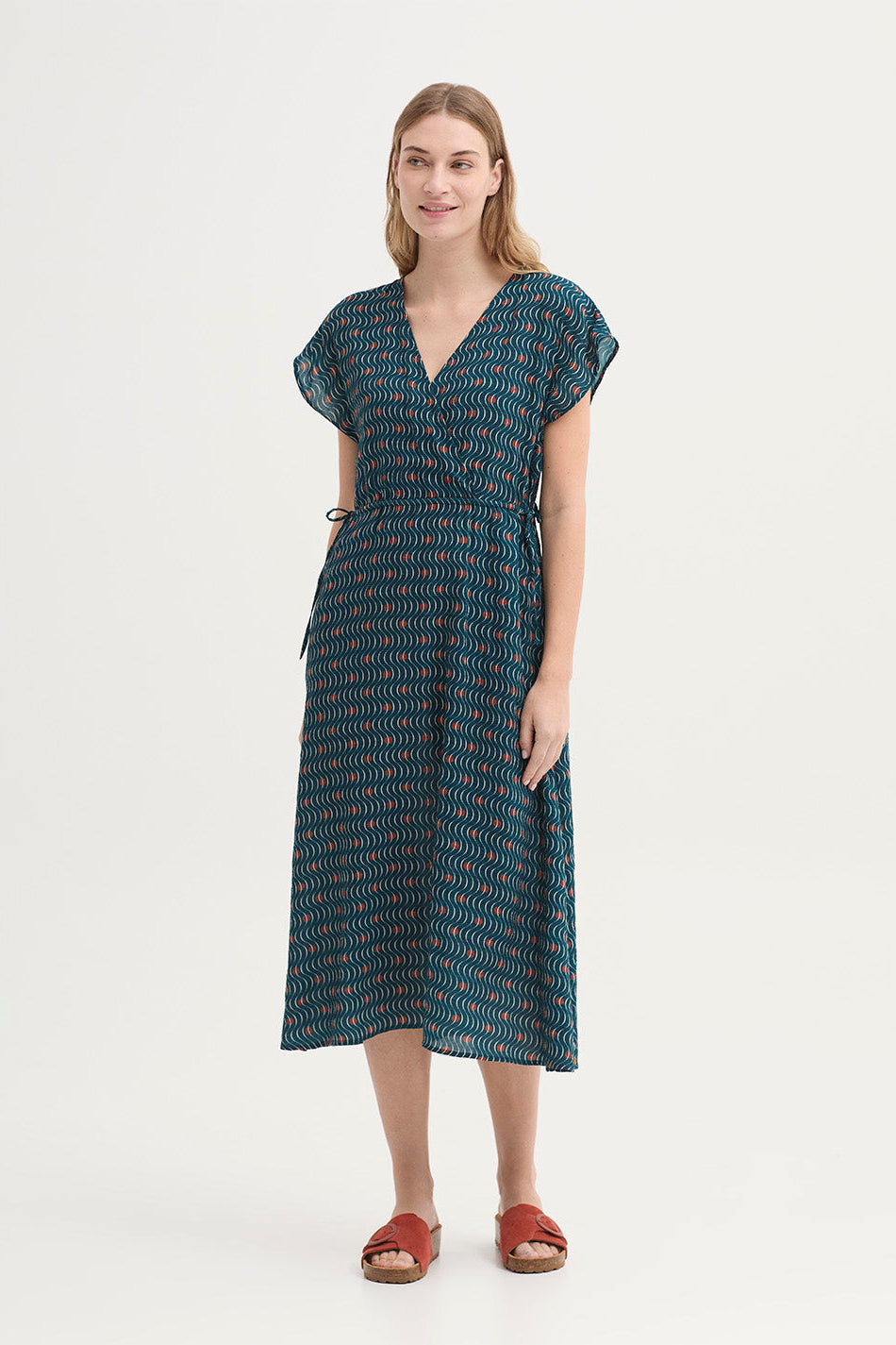 A woman wearing a dark teal wrap-style dress with a wavy abstract pattern in white and rust orange, photographed against a plain light background.