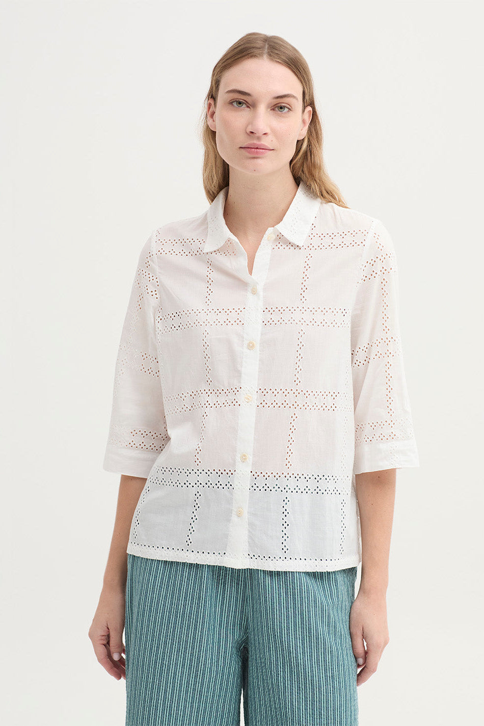 A woman modeling a white eyelet button-up shirt with three-quarter sleeves and green striped trousers, shot against a plain light background.