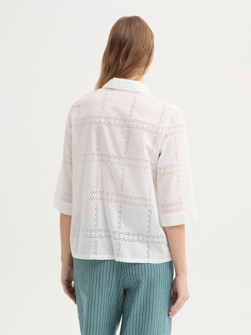 A woman shown from the back modelling a white eyelet button-up shirt with three-quarter sleeves and green striped trousers, shot against a plain light background.