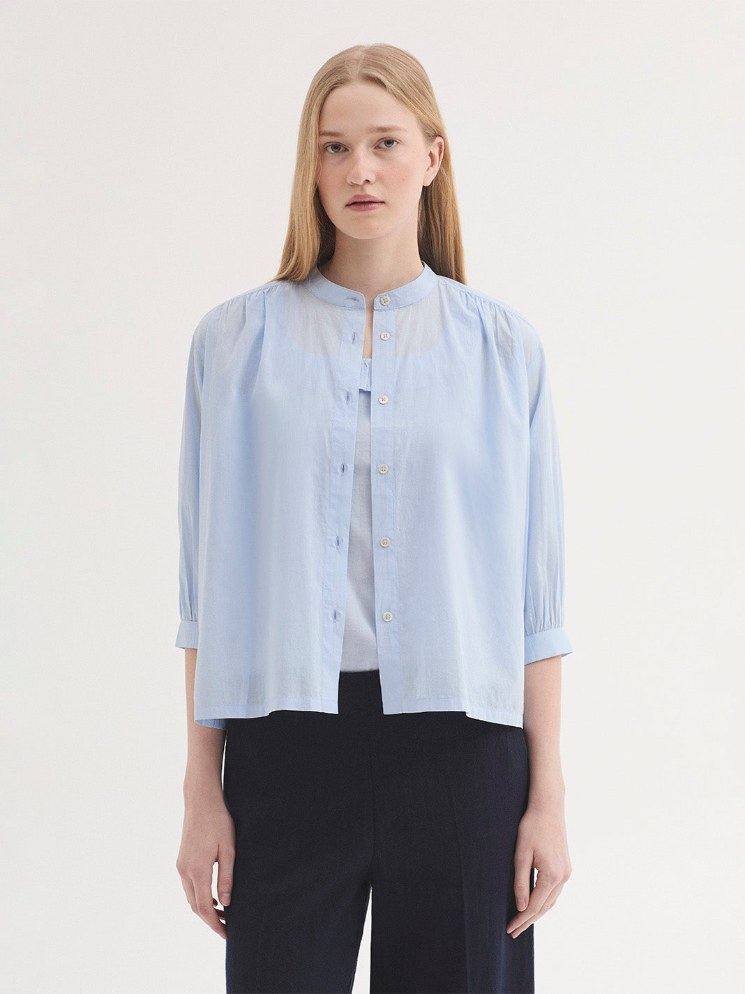 A woman modeling a light blue cropped button-up blouse with a stand collar and 3/4 sleeves, paired with dark navy trousers, against a plain off-white studio background.