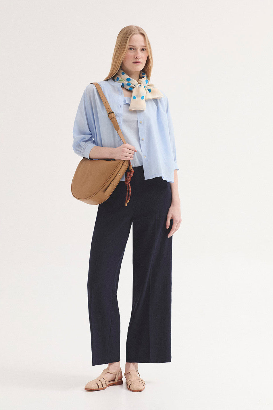 A woman stands against a plain light background wearing a light blue button-up shirt, dark navy wide-leg trousers, a tan shoulder bag, and a patterned blue and yellow neck scarf.