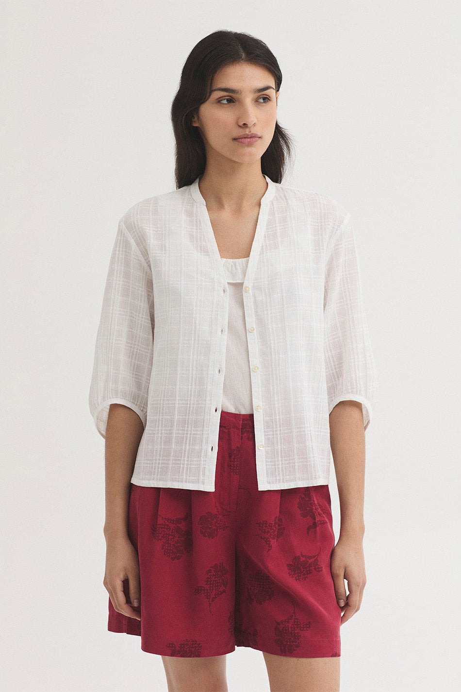 A woman modeling a white textured short-sleeve button-up shirt over a white top, paired with red floral shorts, against a plain light grey studio background.