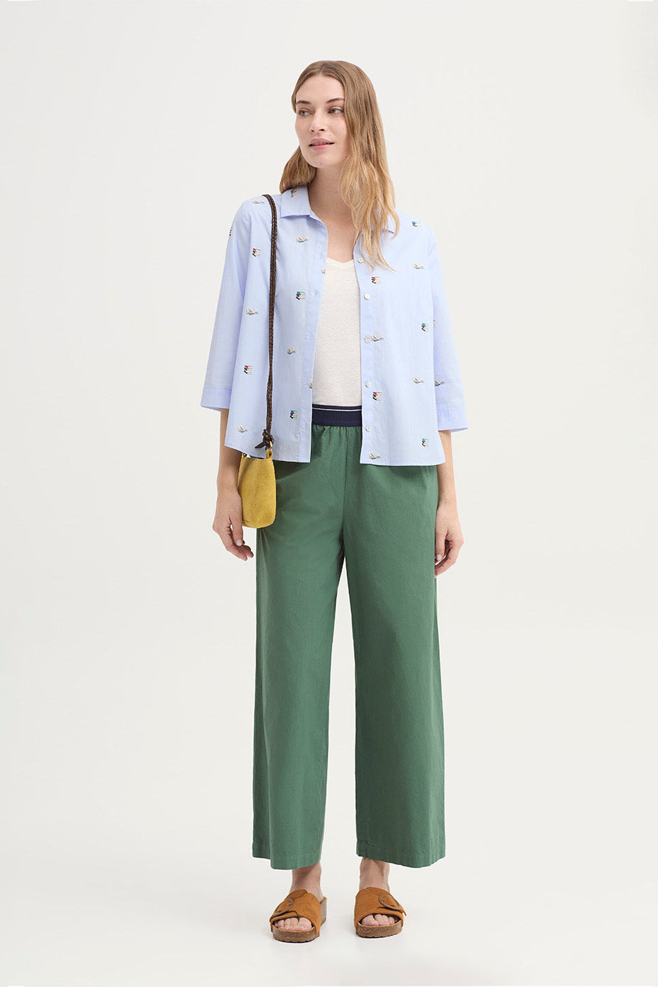 Woman wearing a light blue 3/4 sleeve shirt with delicate books print and mother of pearl buttons with green pants on a white background