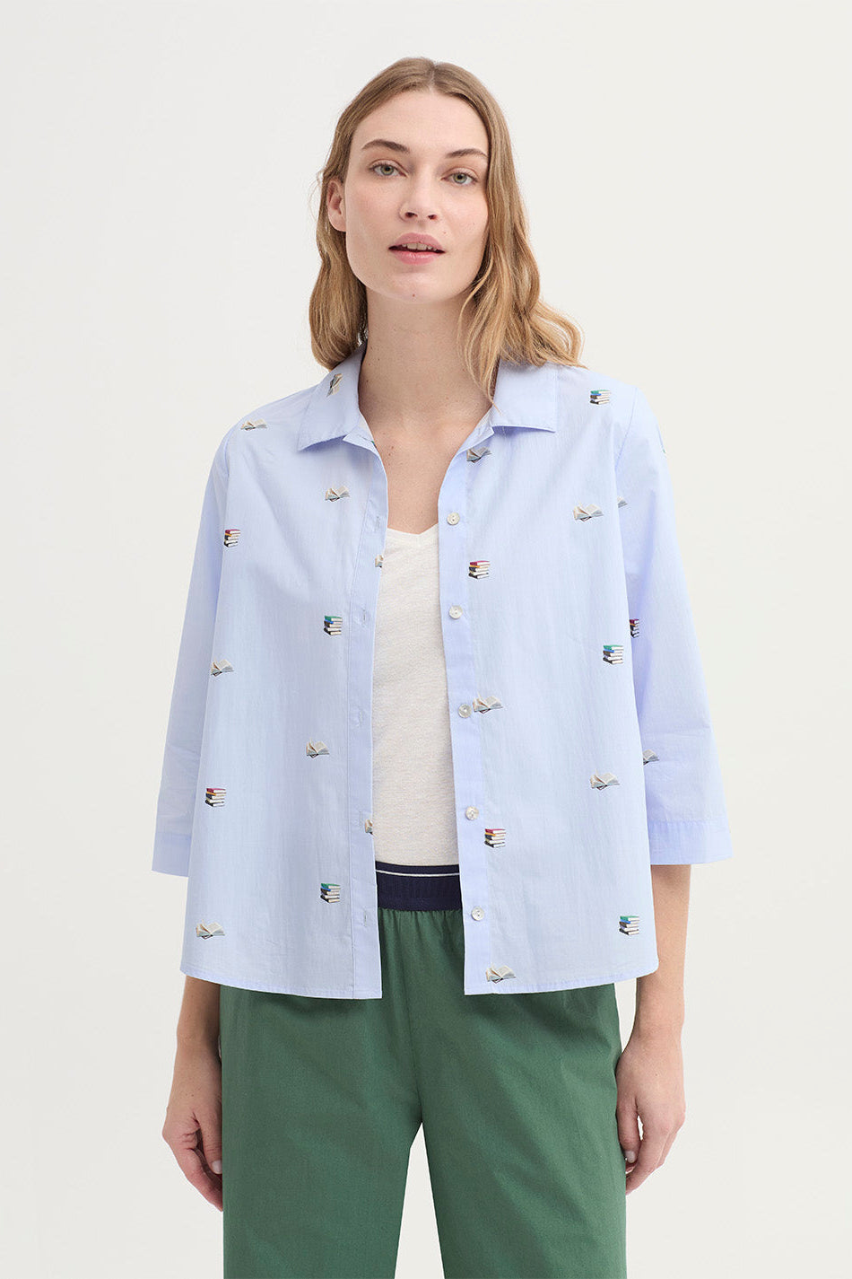 Woman wearing a light blue 3/4 sleeve shirt with delicate books print and mother of pearl buttons with green pants on a white background