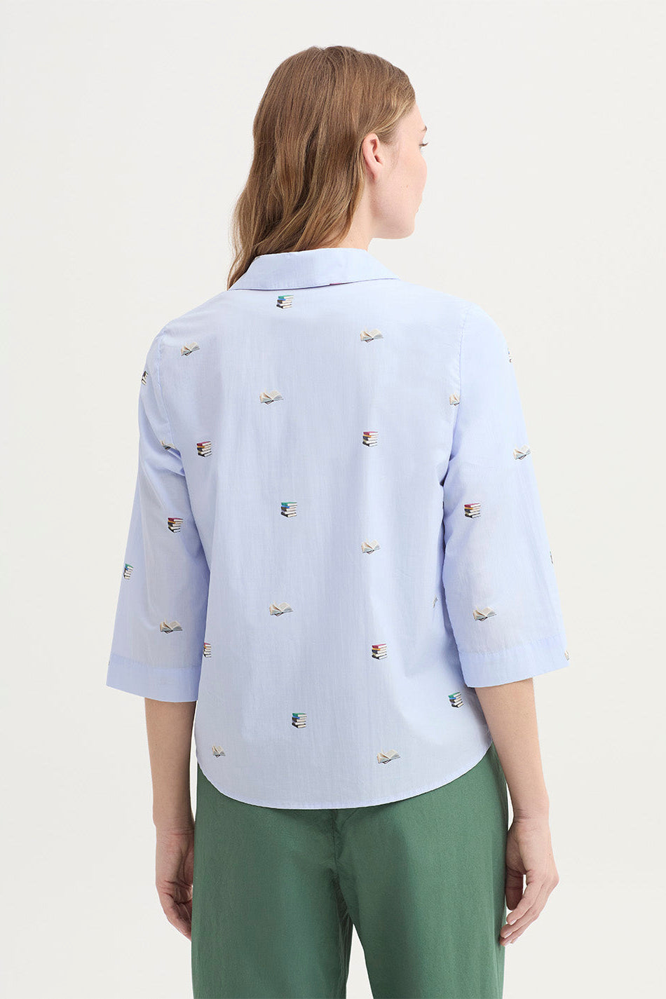 Woman shown from the back wearing a light blue 3/4 sleeve shirt with delicate books print and mother of pearl buttons with green pants on a white background