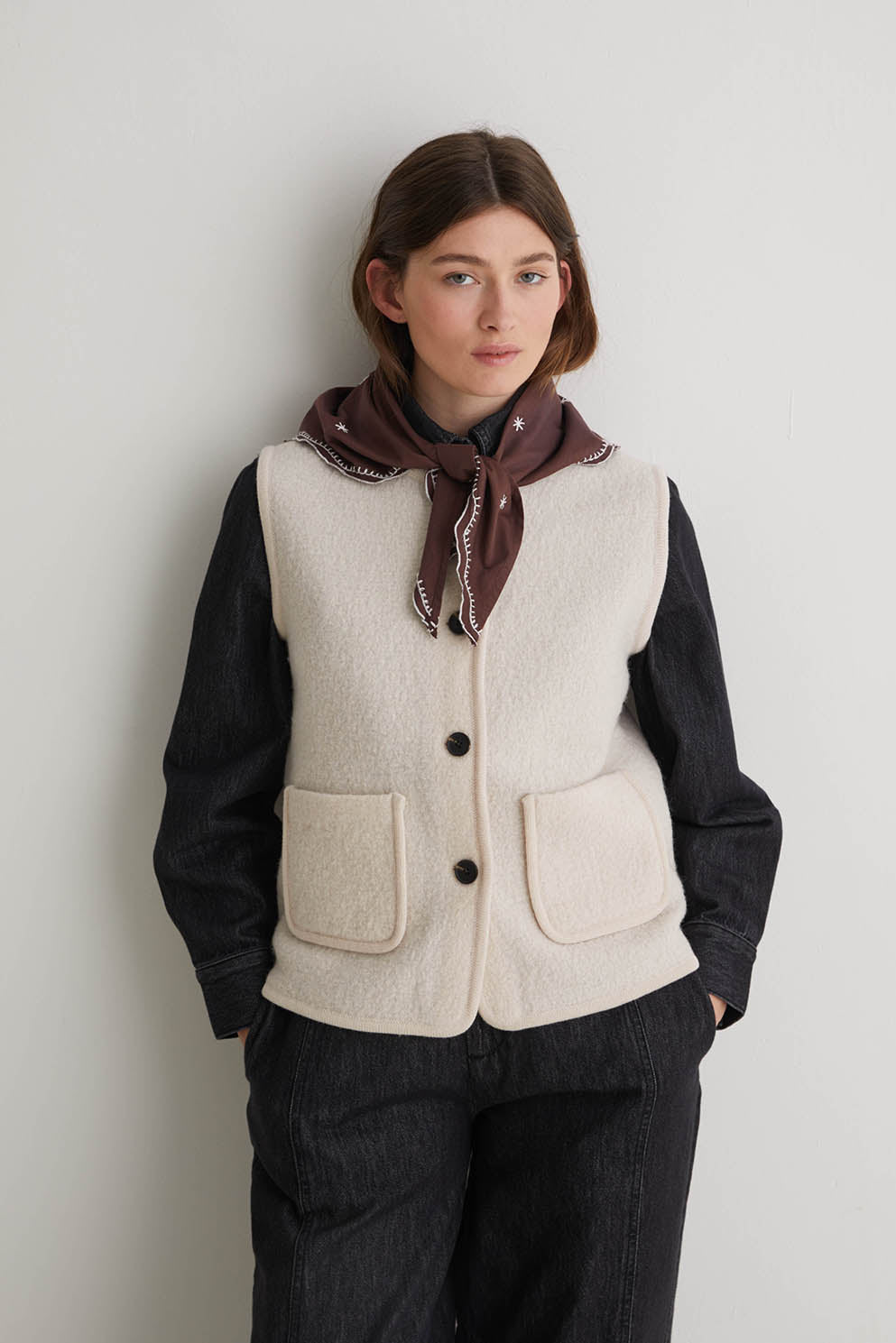 Person wearing a beige vest over a dark shirt with a scarf, against a plain background