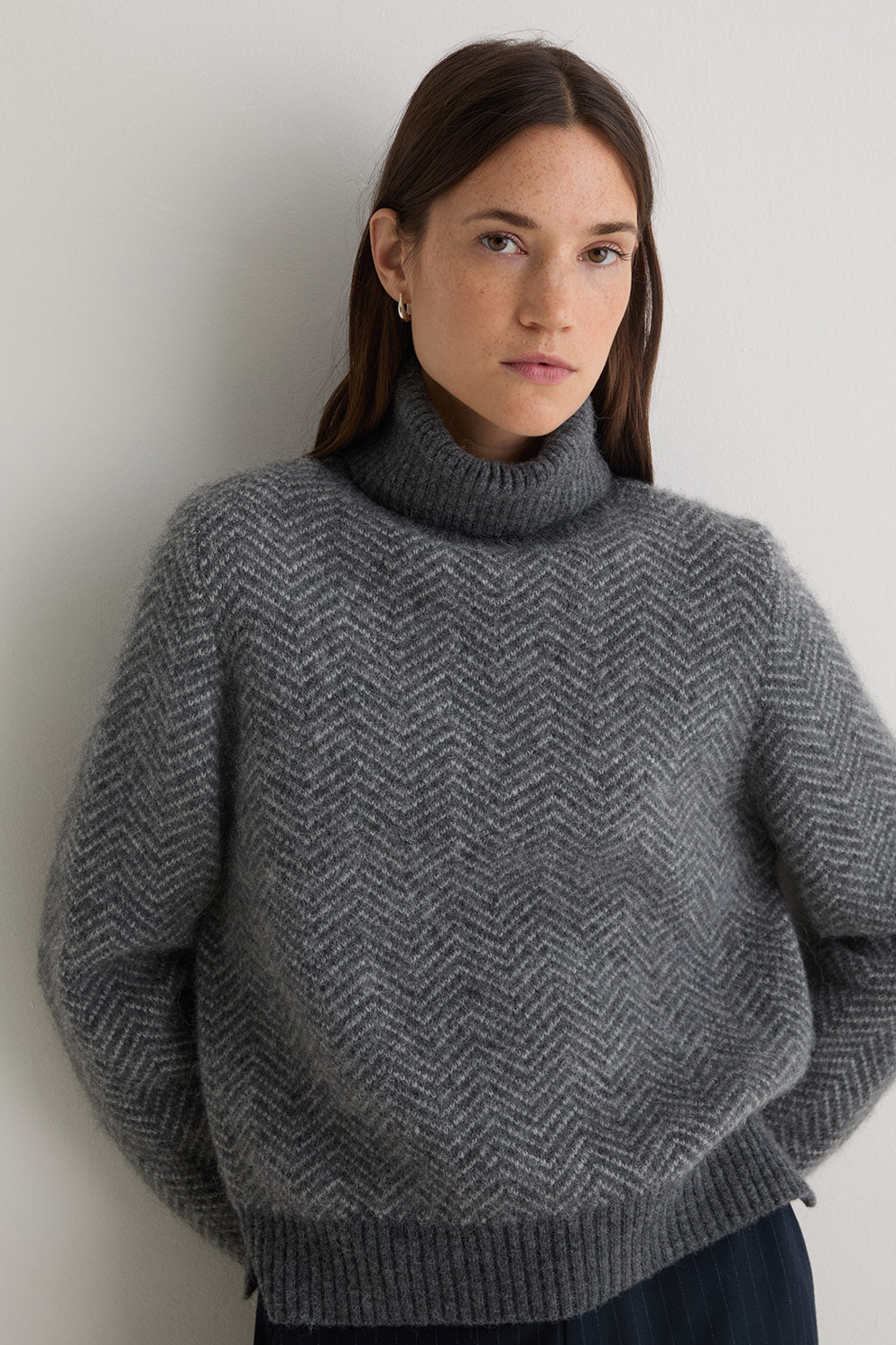 Person wearing a grey turtle neck herringbone sweater and black pants on a white background
