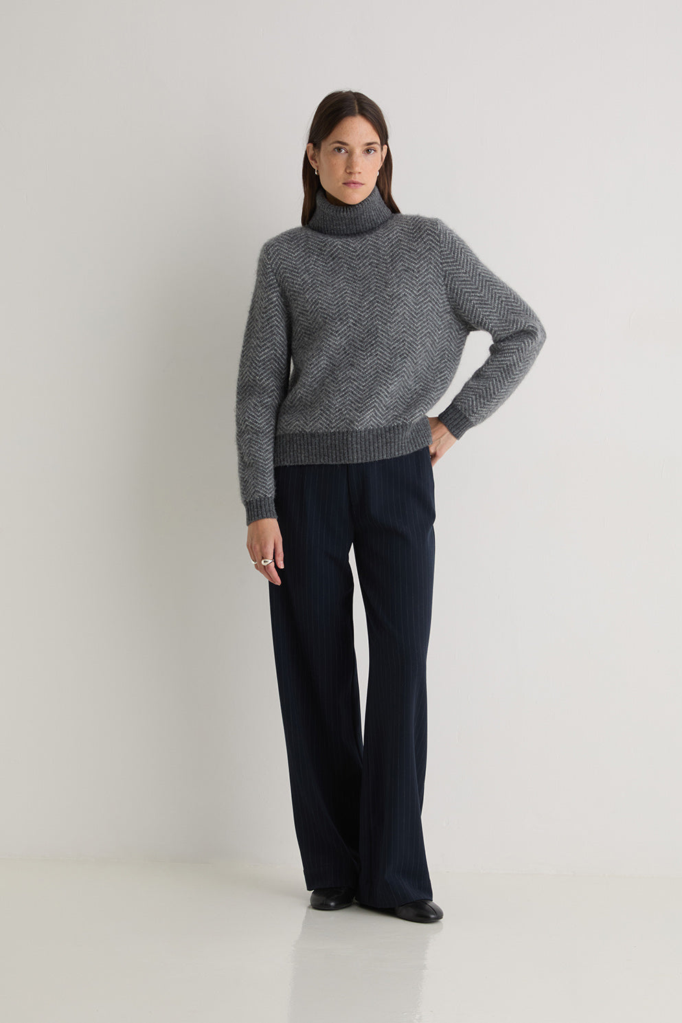 Person wearing a grey turtle neck herringbone sweater and black pants on a white background