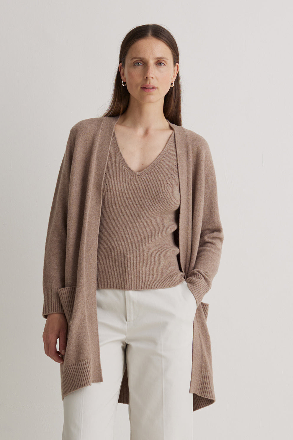 Woman wearing a brown cardigan over a beige top and white pants on a light gray background