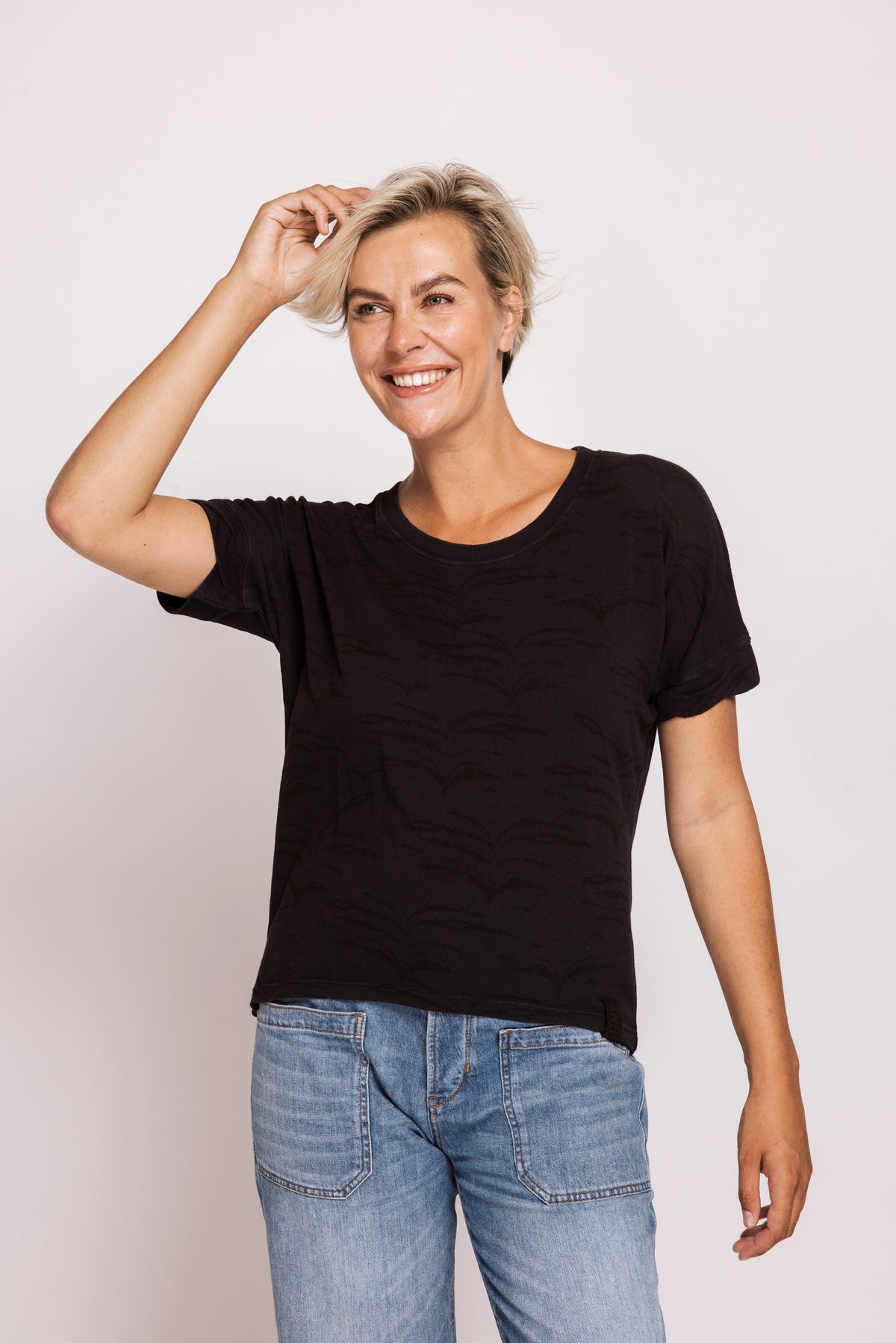 A smiling woman with short blonde hair wearing a black short-sleeve crewneck t-shirt and light blue jeans, posing against a light gray studio background.