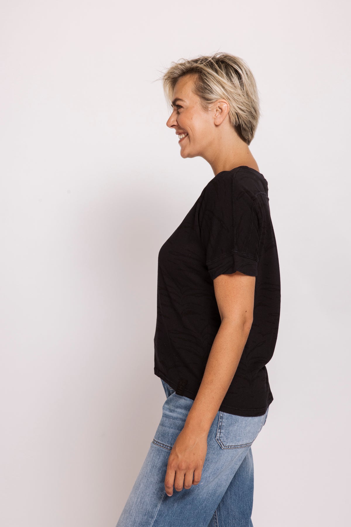 A woman with short blonde hair is shown in profile wearing a black short-sleeve t-shirt and light blue jeans against a plain white studio background.
