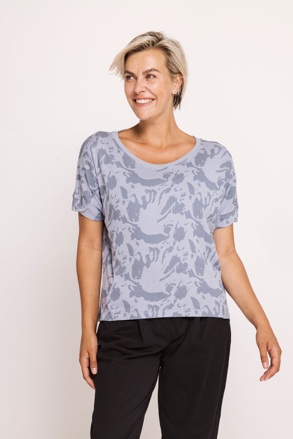 A woman modeling a short-sleeve scoop-neck top with a light blue and gray abstract pattern, paired with black pants, against a plain white studio background.