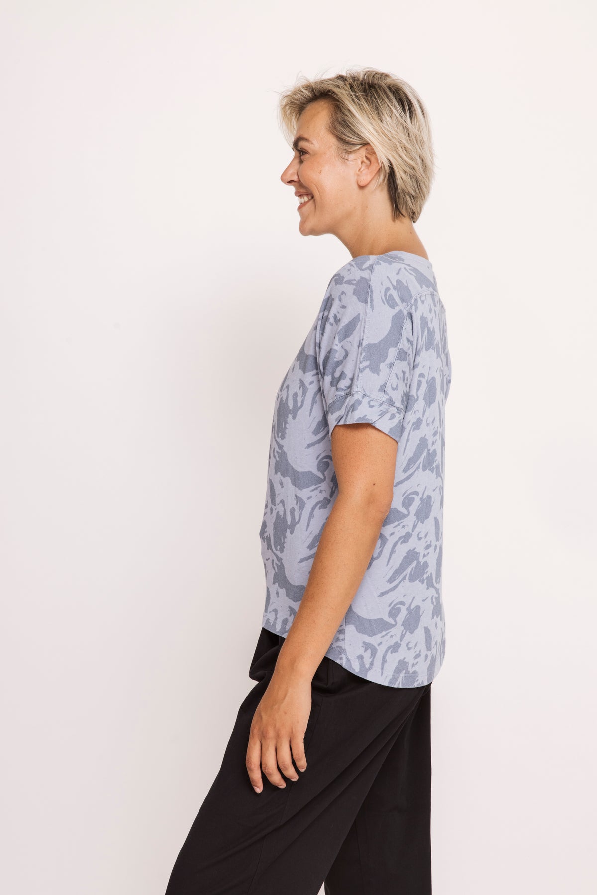 A woman with short blonde hair is shown in profile wearing a light blue-gray short-sleeve top with a darker blue-gray abstract pattern, paired with black pants, against a plain off-white background.