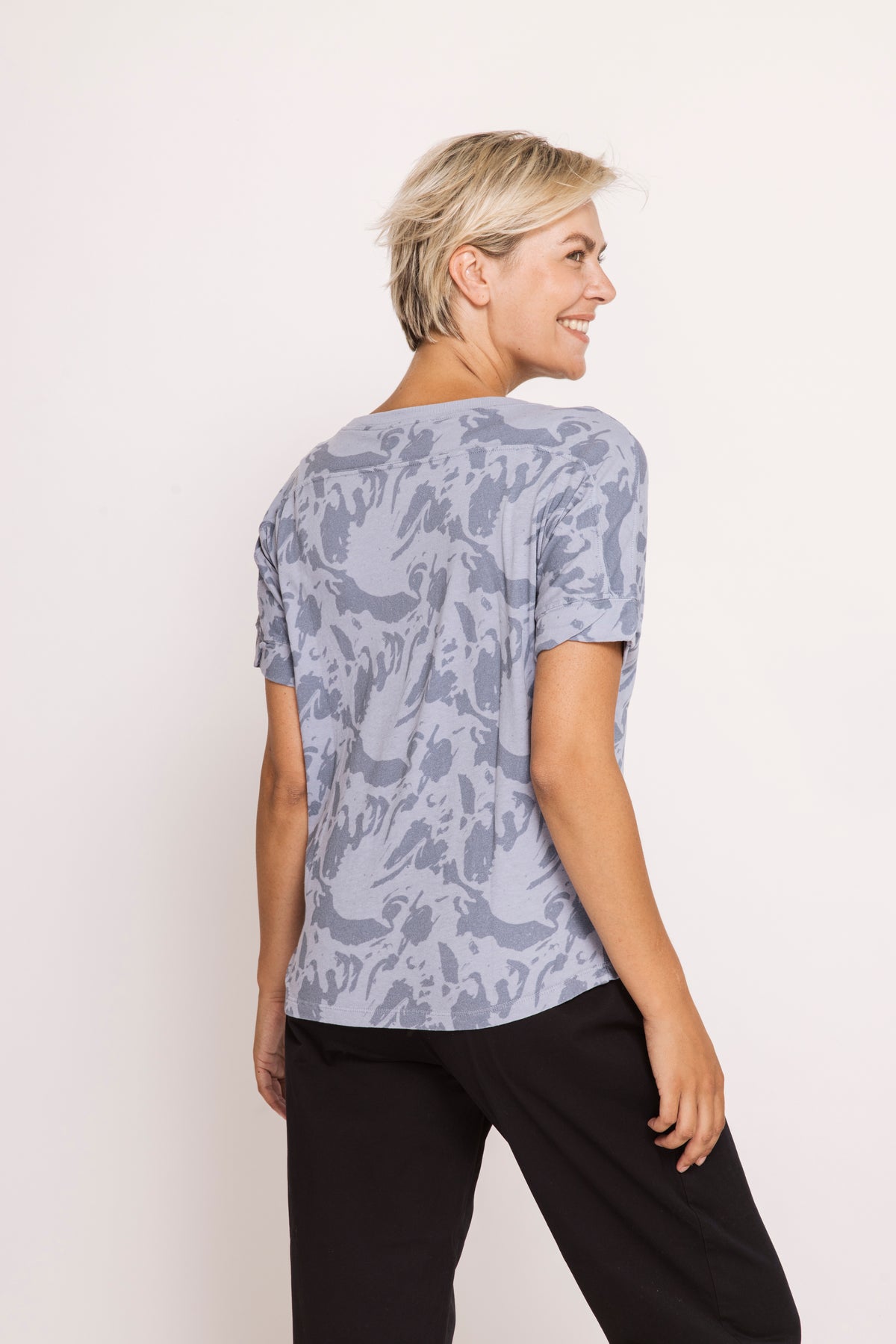 A woman with short blonde hair is shown from the back wearing a light blue-gray short-sleeve t-shirt with a darker gray abstract pattern, paired with black pants, against a plain off-white background.