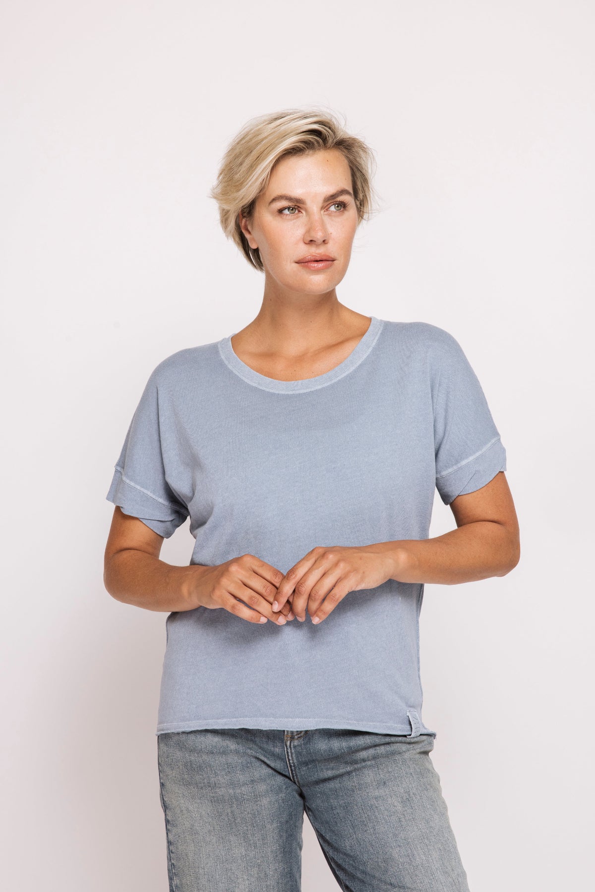 A woman with short blonde hair wearing a light blue short-sleeve knit top and blue jeans, standing against a plain light gray studio background.