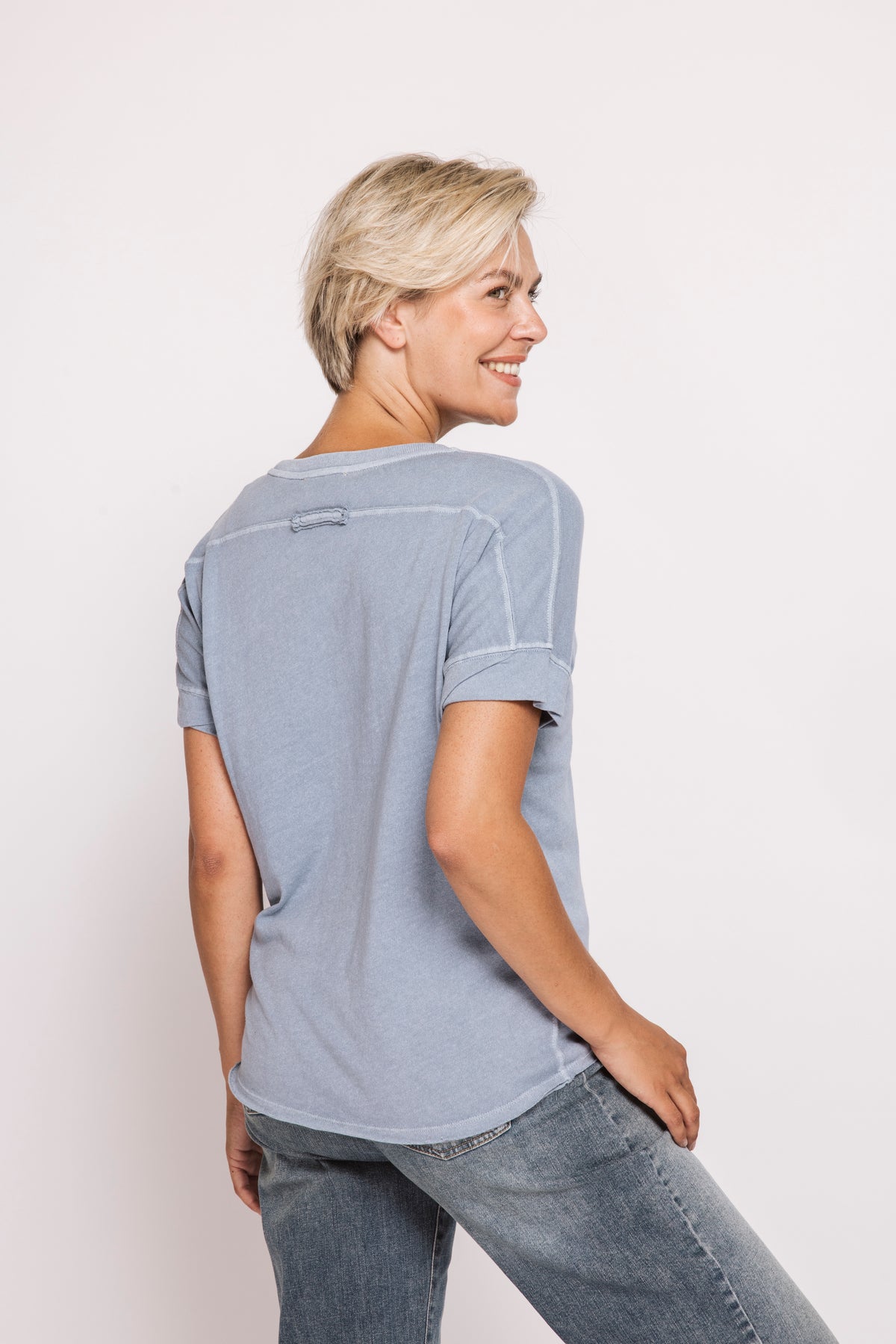 A woman with short blonde hair is shown from the back wearing a light blue-gray short-sleeve t-shirt with contrast stitching and blue jeans, set against a plain off-white studio background.