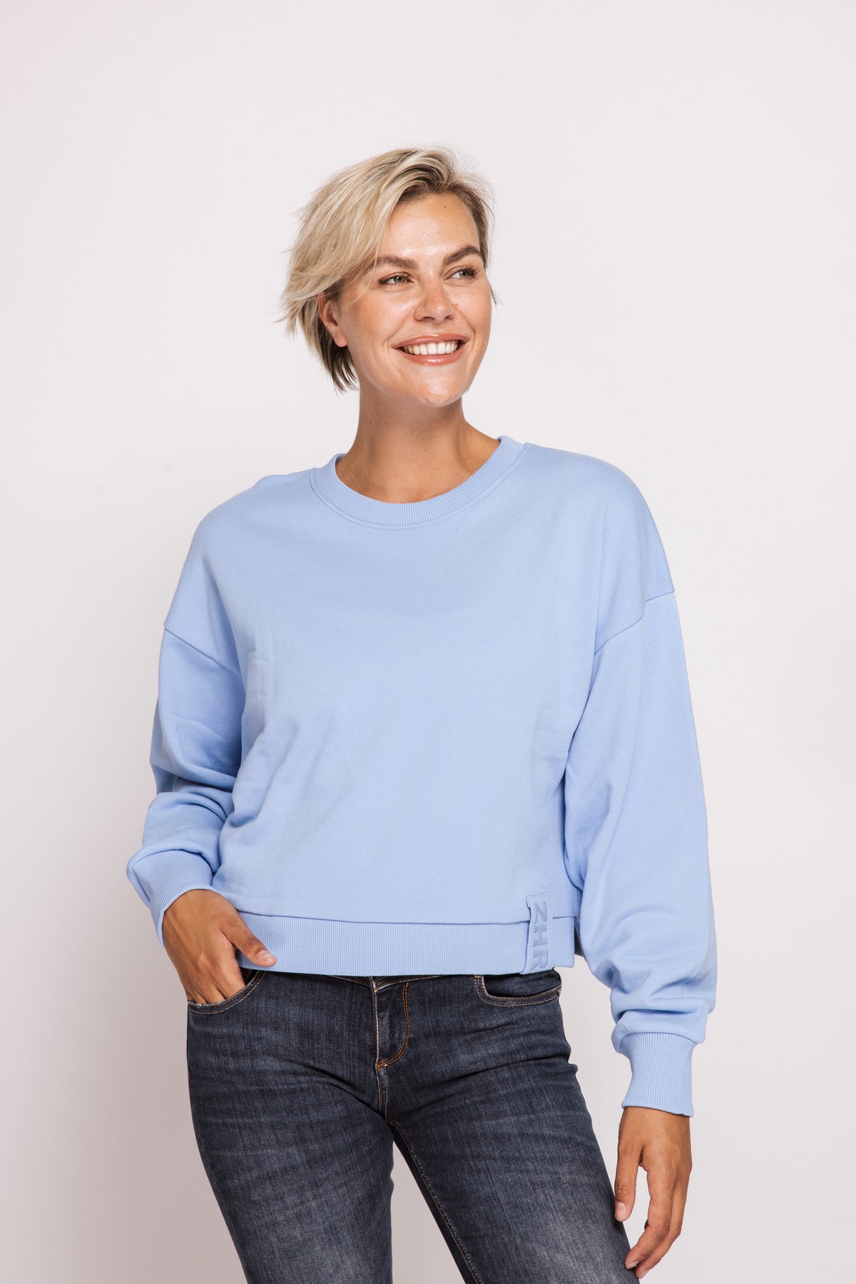 A woman with short blonde hair wearing a light blue crewneck sweatshirt and dark blue jeans, standing against a plain light gray studio background.