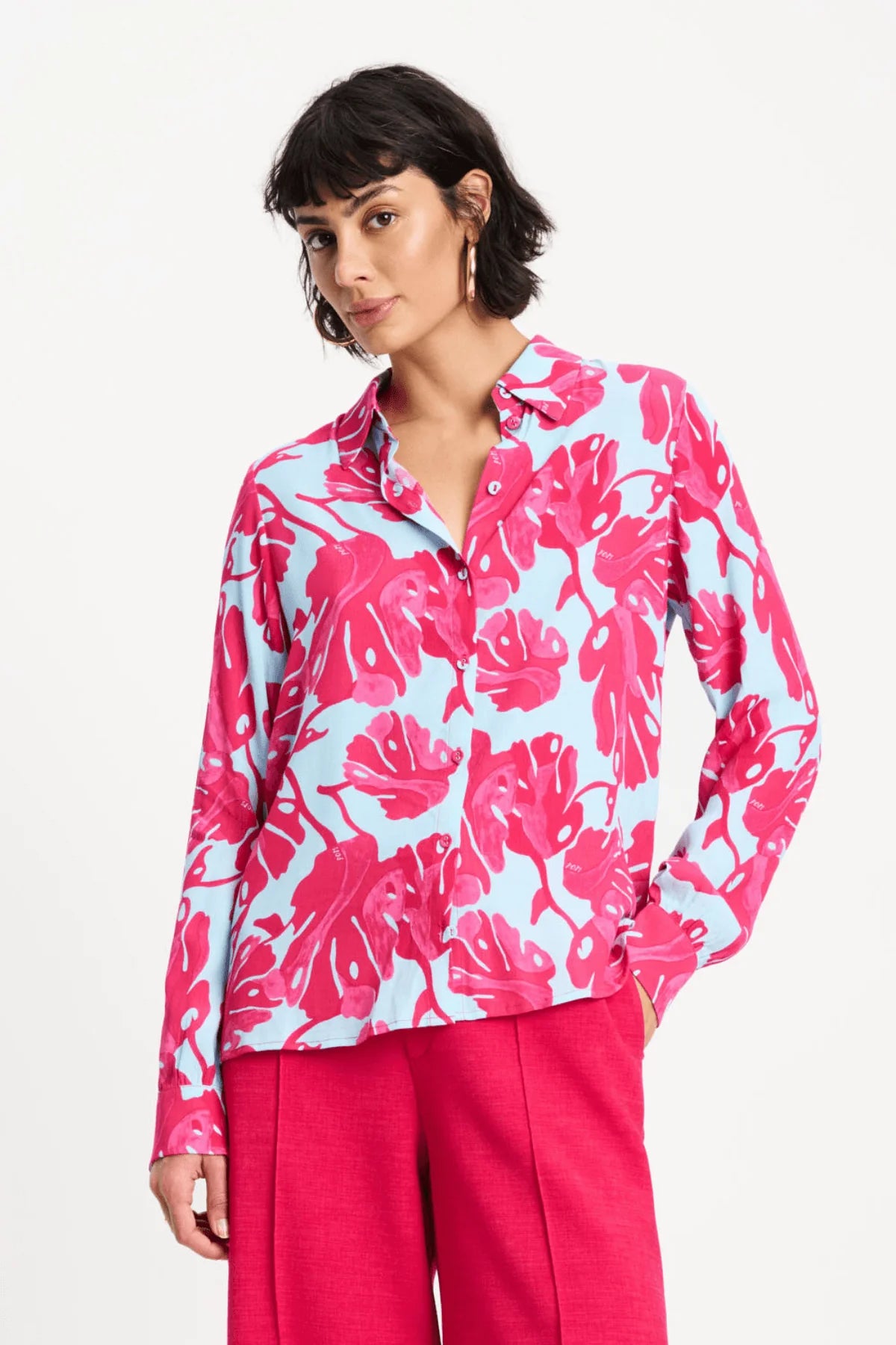 A woman modeling a long-sleeve button-up shirt with a bold pink tropical leaf print on a light blue background, paired with bright pink trousers, against a plain white studio background.