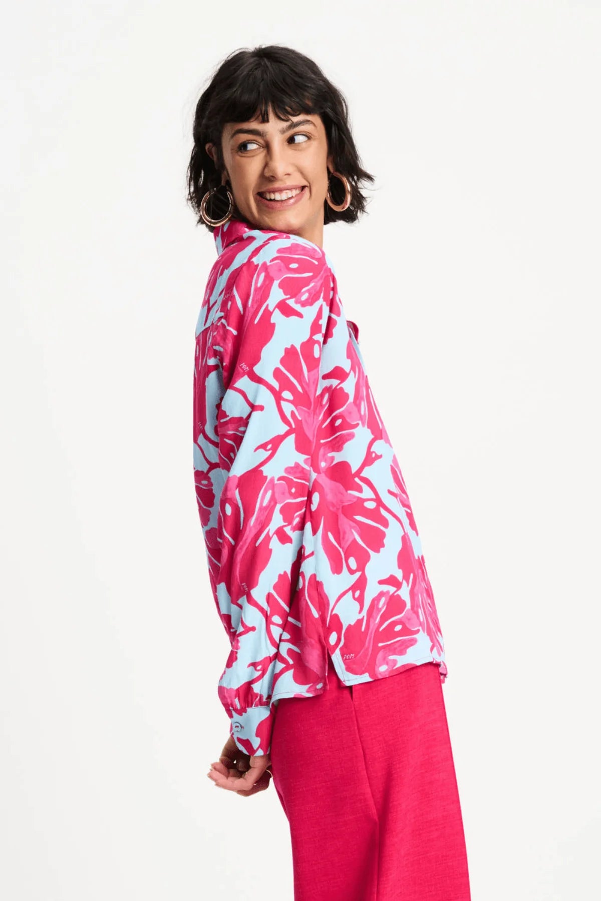 A woman modeling a long-sleeve button-up shirt with a bold pink and light blue tropical floral pattern, paired with bright pink pants, against a plain white studio background.