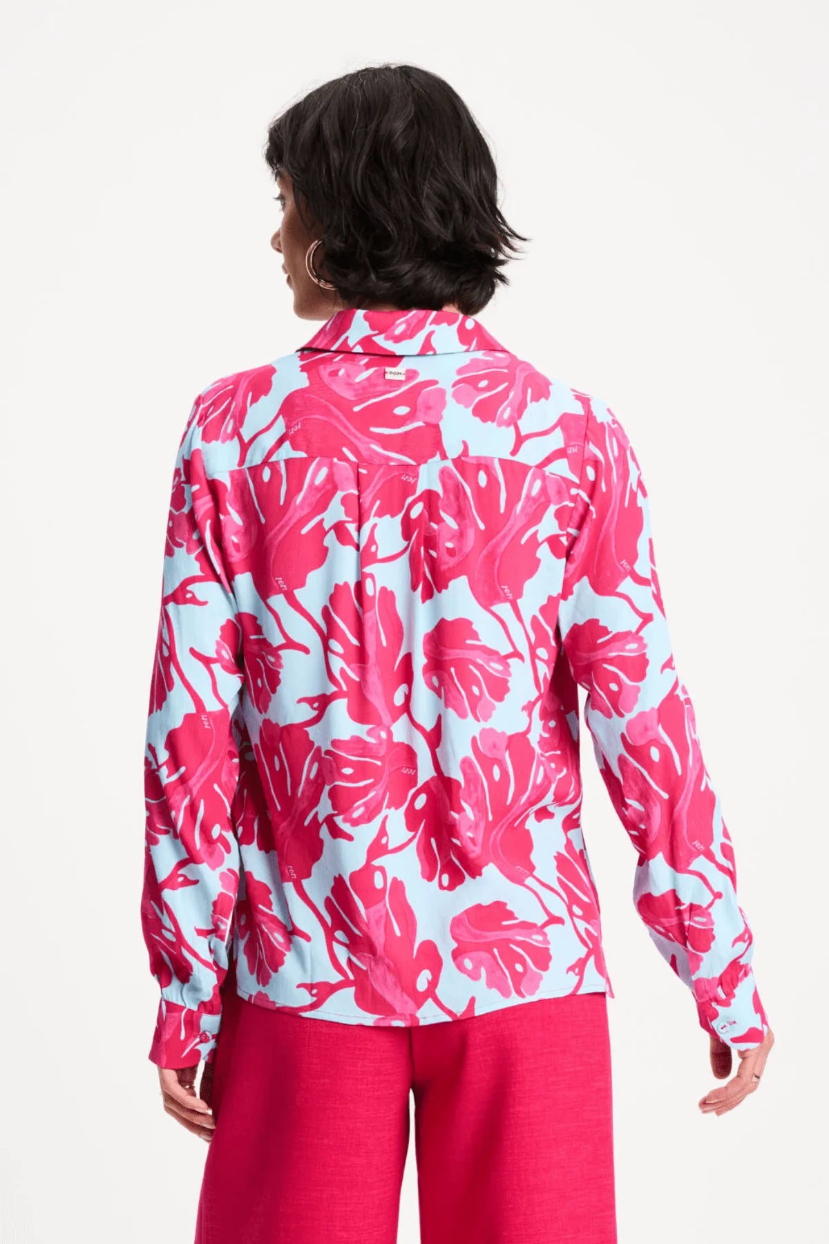 A woman shown from the back wearing a light blue long-sleeve button-up shirt with a bold hot pink tropical floral print, paired with hot pink pants, against a plain white studio background.