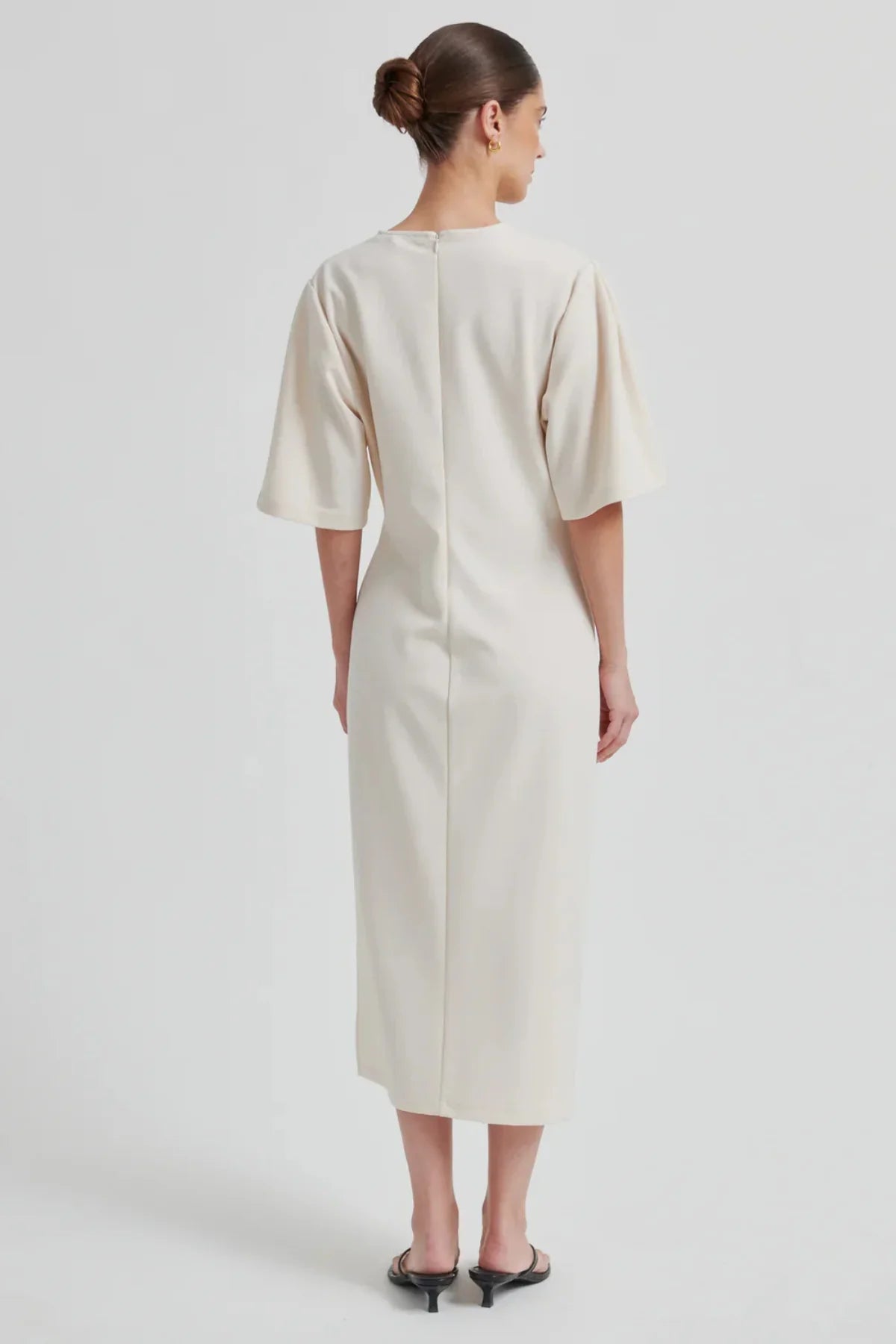 A women shown from the back wearing a midi length beige dress with shorts sleeves and black heels on a white background.  