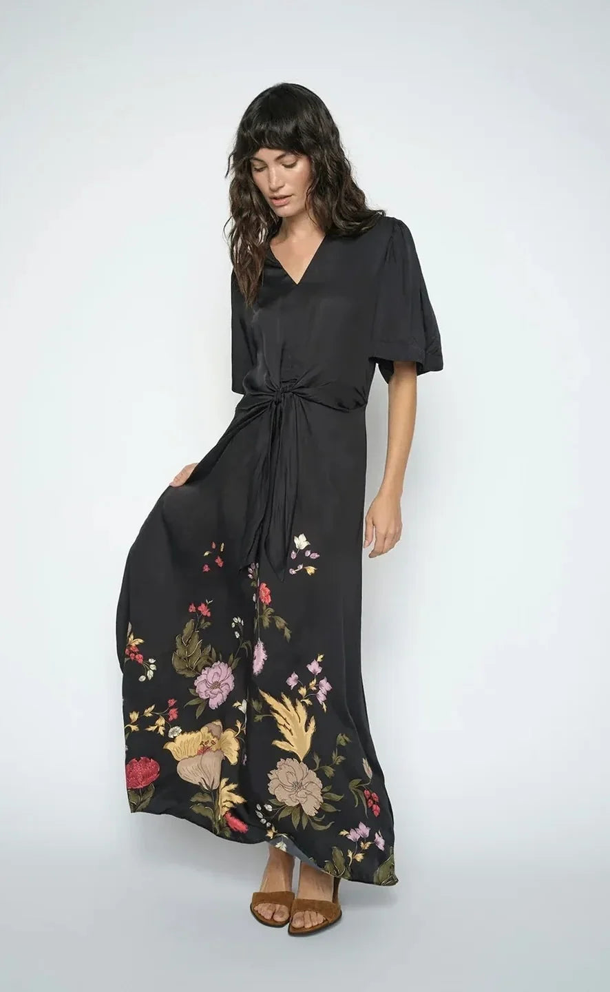 A women  wearing a  black wrap-style maxi dress with short flutter sleeves, a V-neckline, a front waist tie, and a floral print concentrated along the lower skirt, shown on a plain white background.