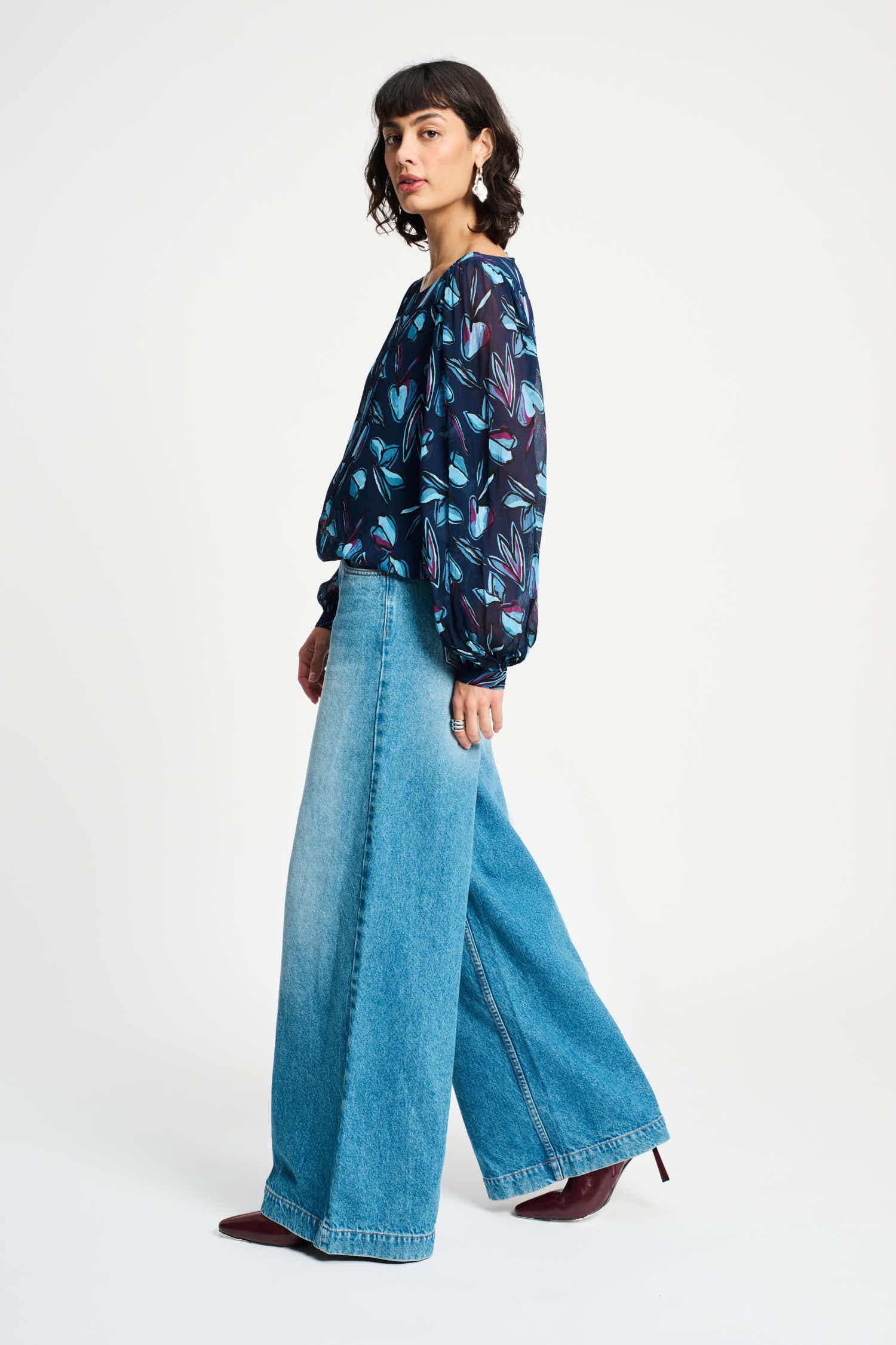 Woman wearing a colourful patterned blouse with long sleeves and light blue jeans on a white background