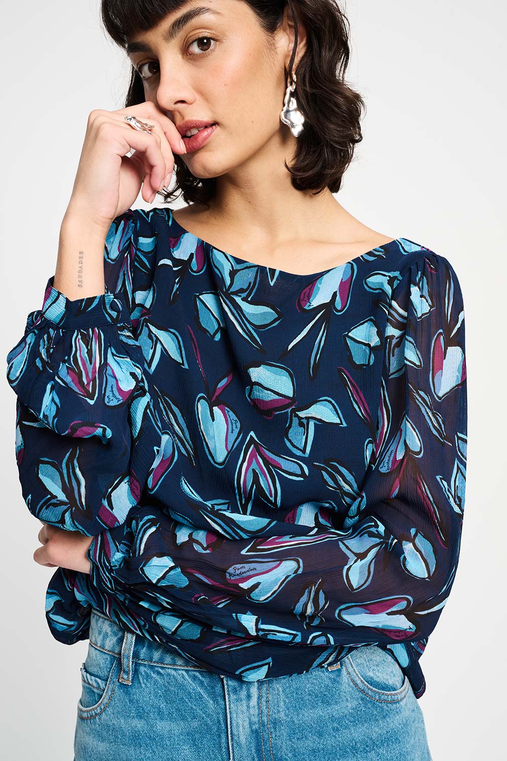 Woman wearing a colourful patterned blouse with long sleeves and light blue jeans on a white background