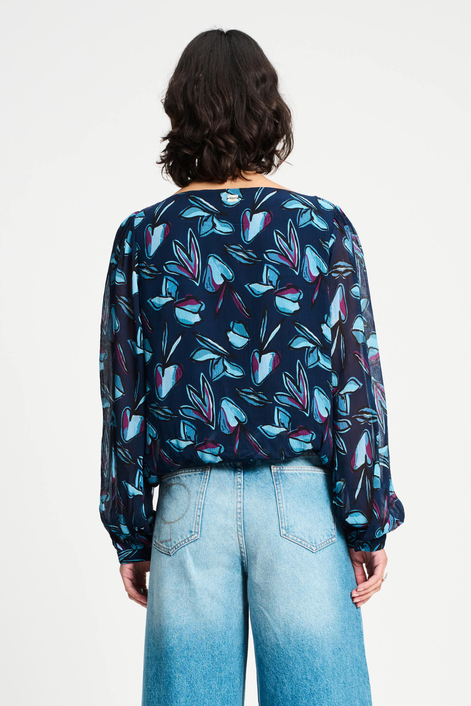 Woman wearing a colourful patterned blouse with long sleeves and light blue jeans on a white background