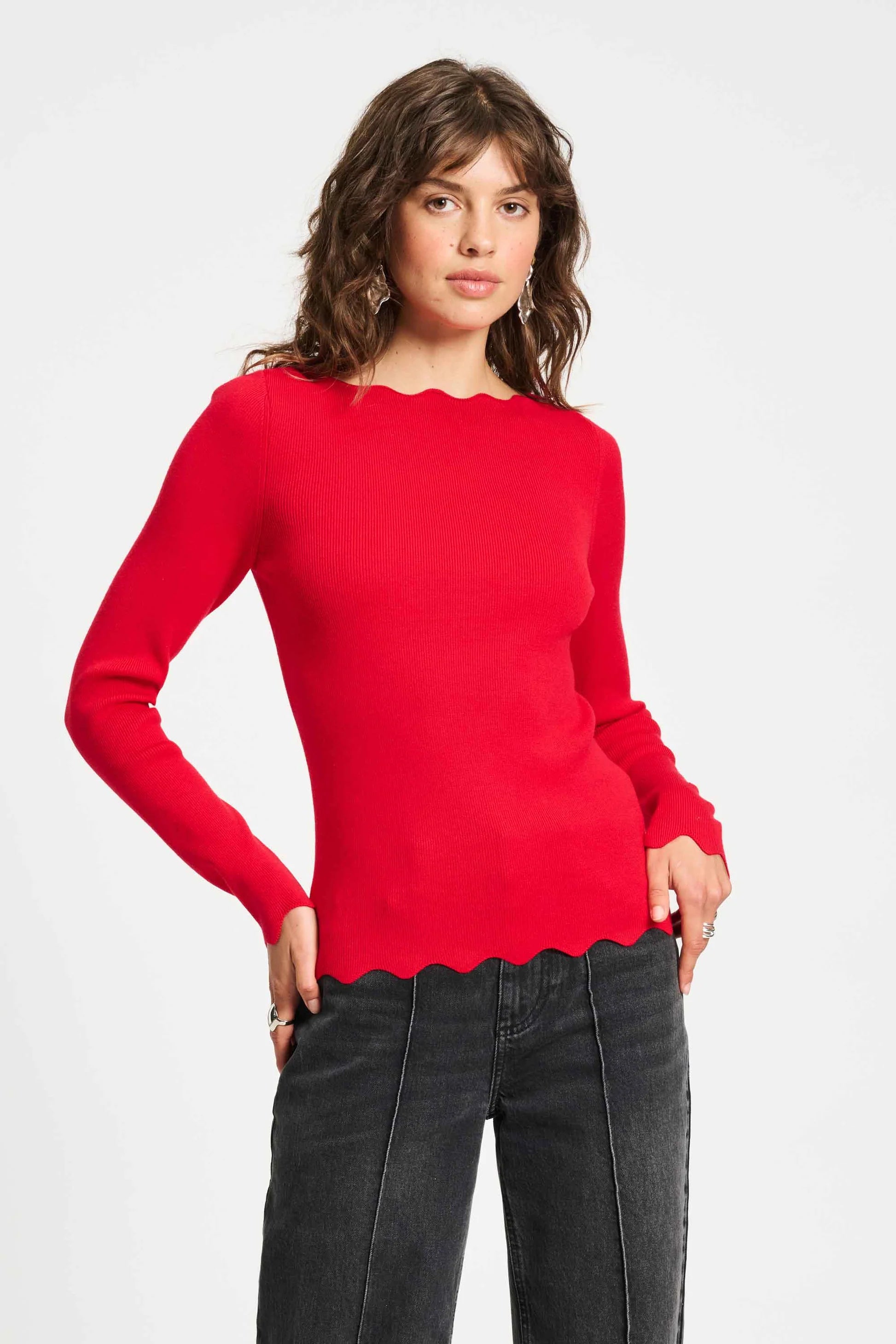 Woman wearing a red sweater with scalloped edges and grey wash jeans on a white background